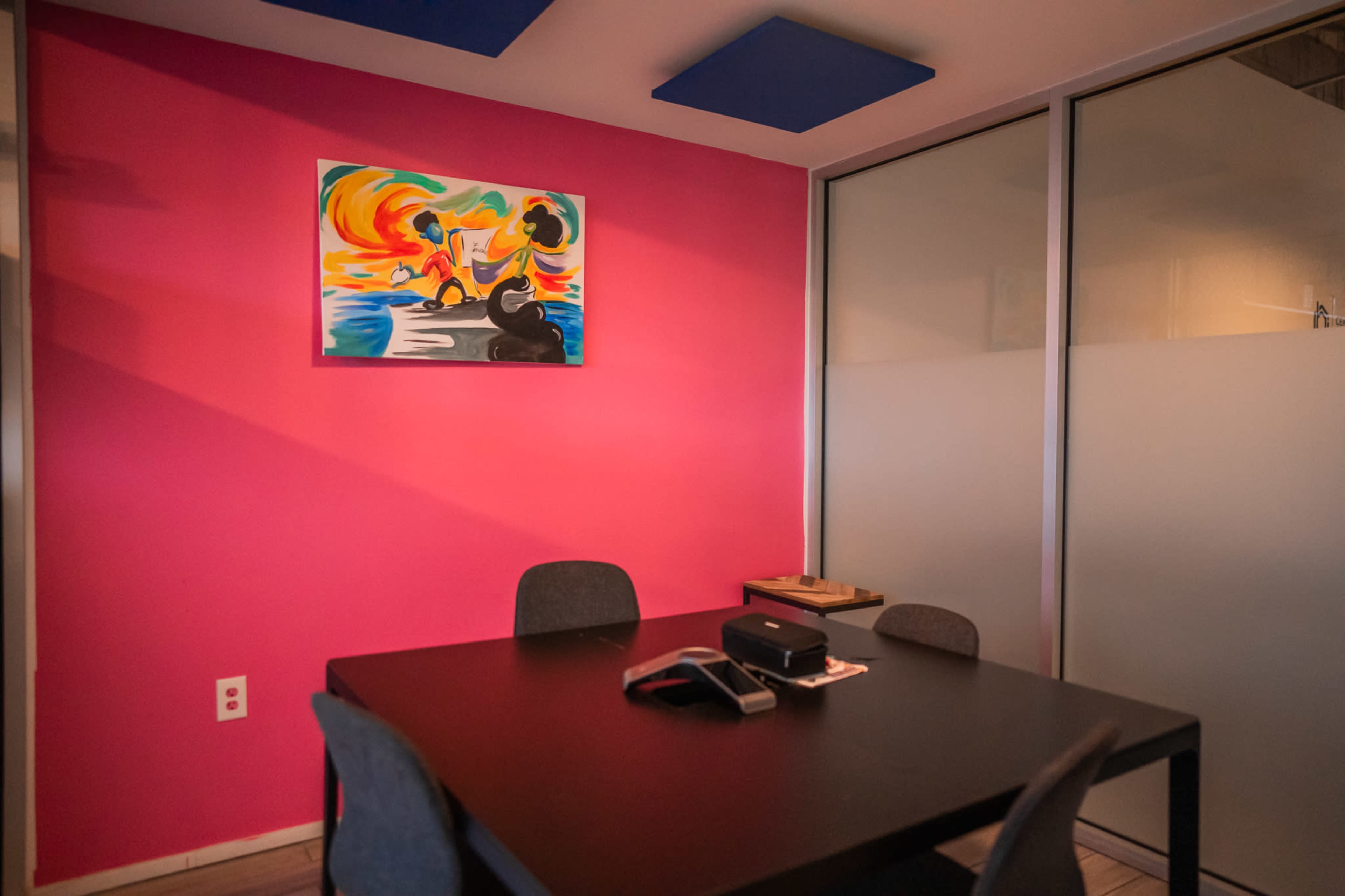 A small meeting room with a pink wall, a black table, and four chairs, featuring a colorful painting on the wall.