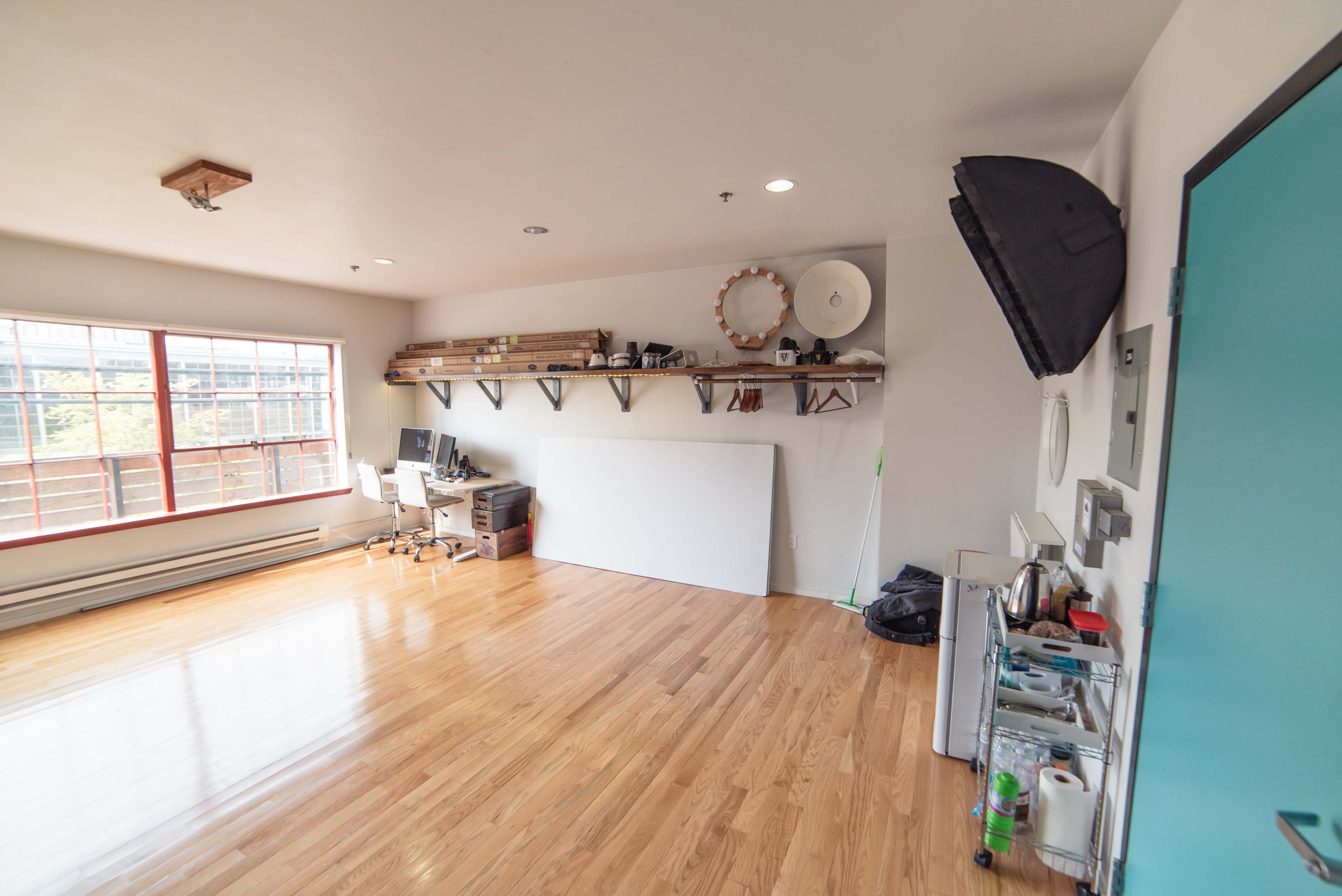 A spacious room with wooden floors, a large window, a desk set up for work, and shelves holding various items along the wall.