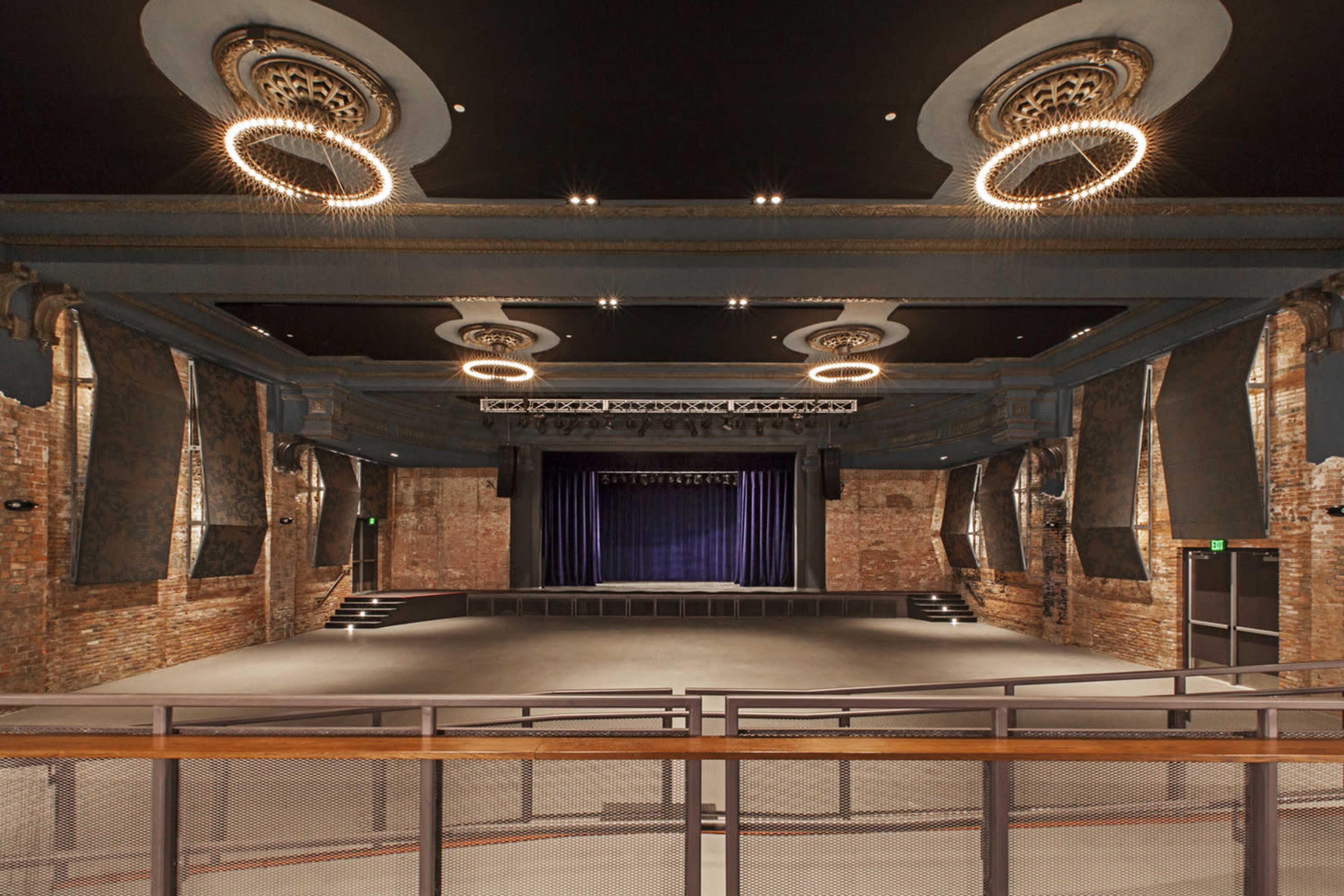 A spacious auditorium with exposed brick walls, a raised stage draped in blue curtains, and decorative lighting fixtures on the ceiling.