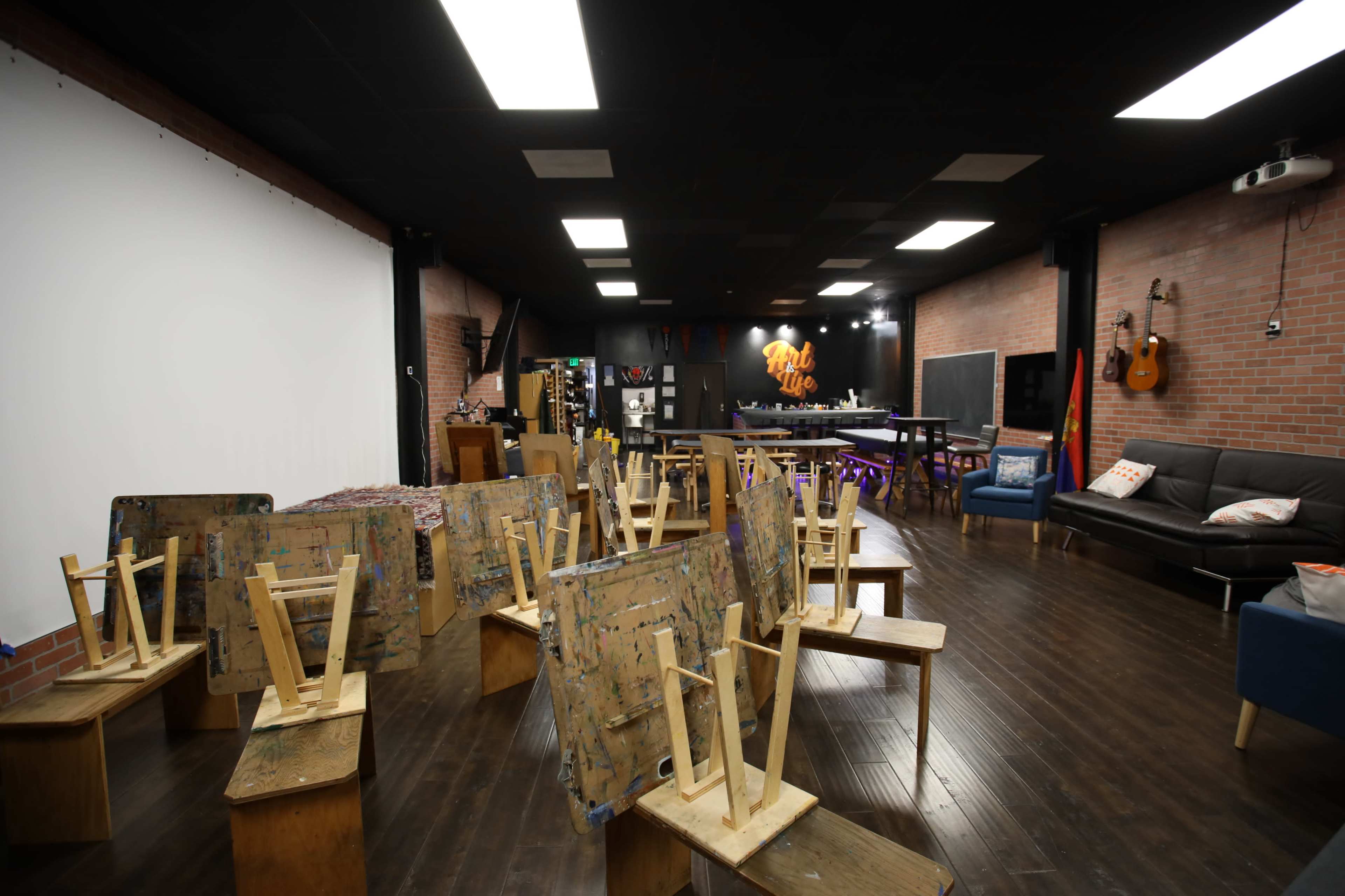 The image shows a large studio space with wooden tables stacked upside down, surrounded by brick walls and a kitchen area in the background.
