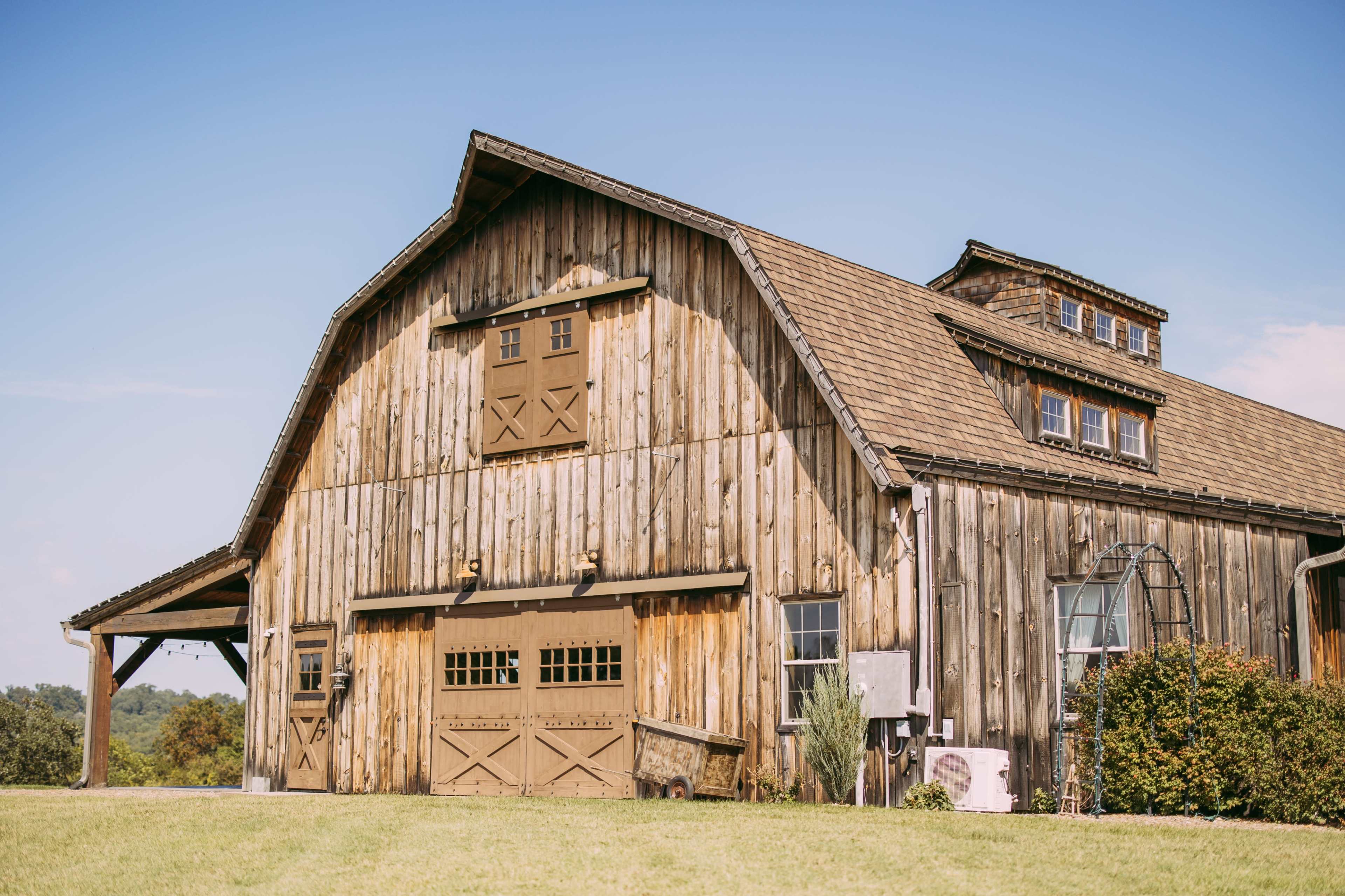 A large wooden barn with a sloped roof and multiple windows sits in a grassy field under a clear blue sky.