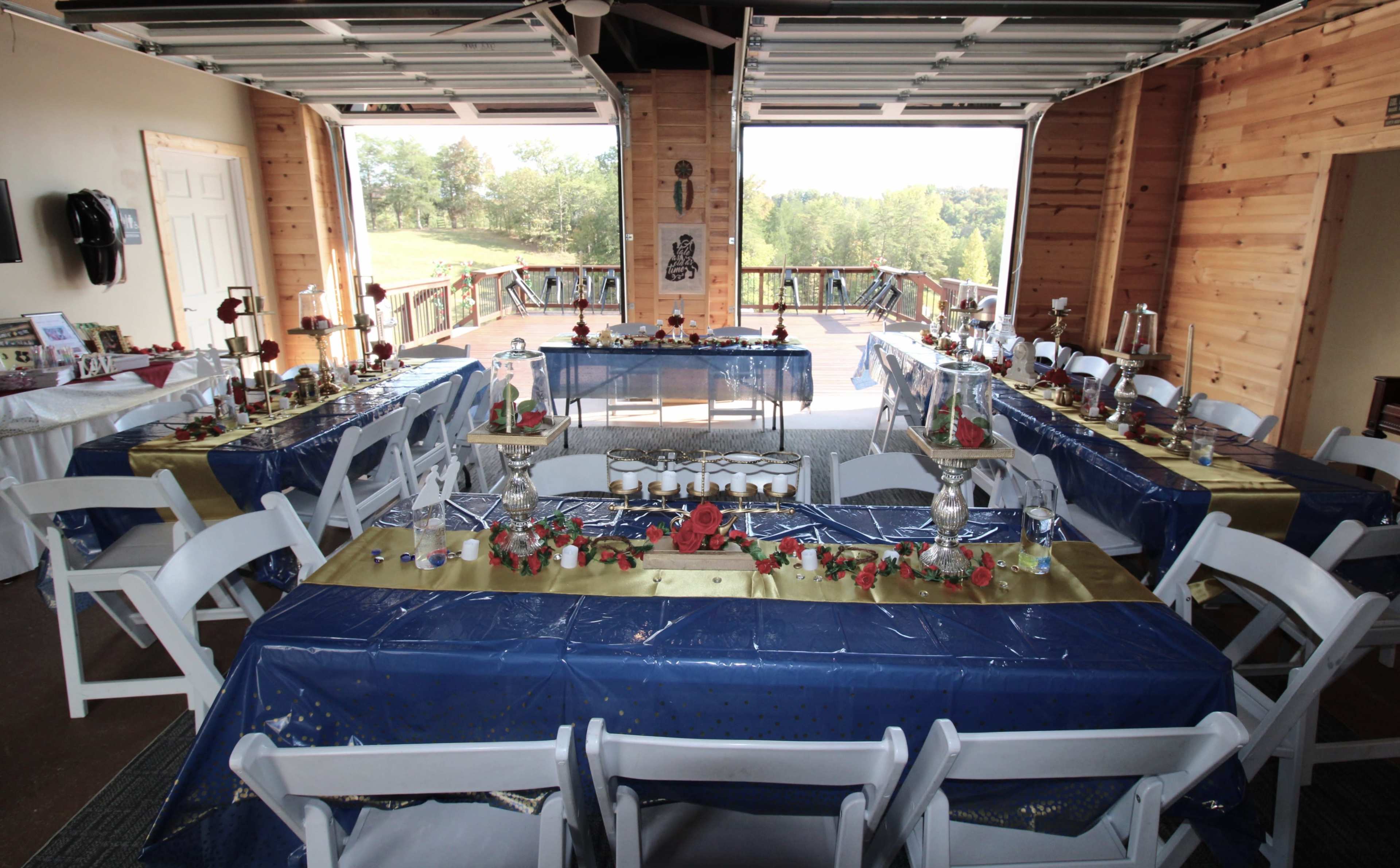 The image shows a decorated indoor venue with several tables covered in blue and gold tablecloths, set with centerpieces and surrounded by white chairs, all facing an open door leading to a scenic outdoor view.