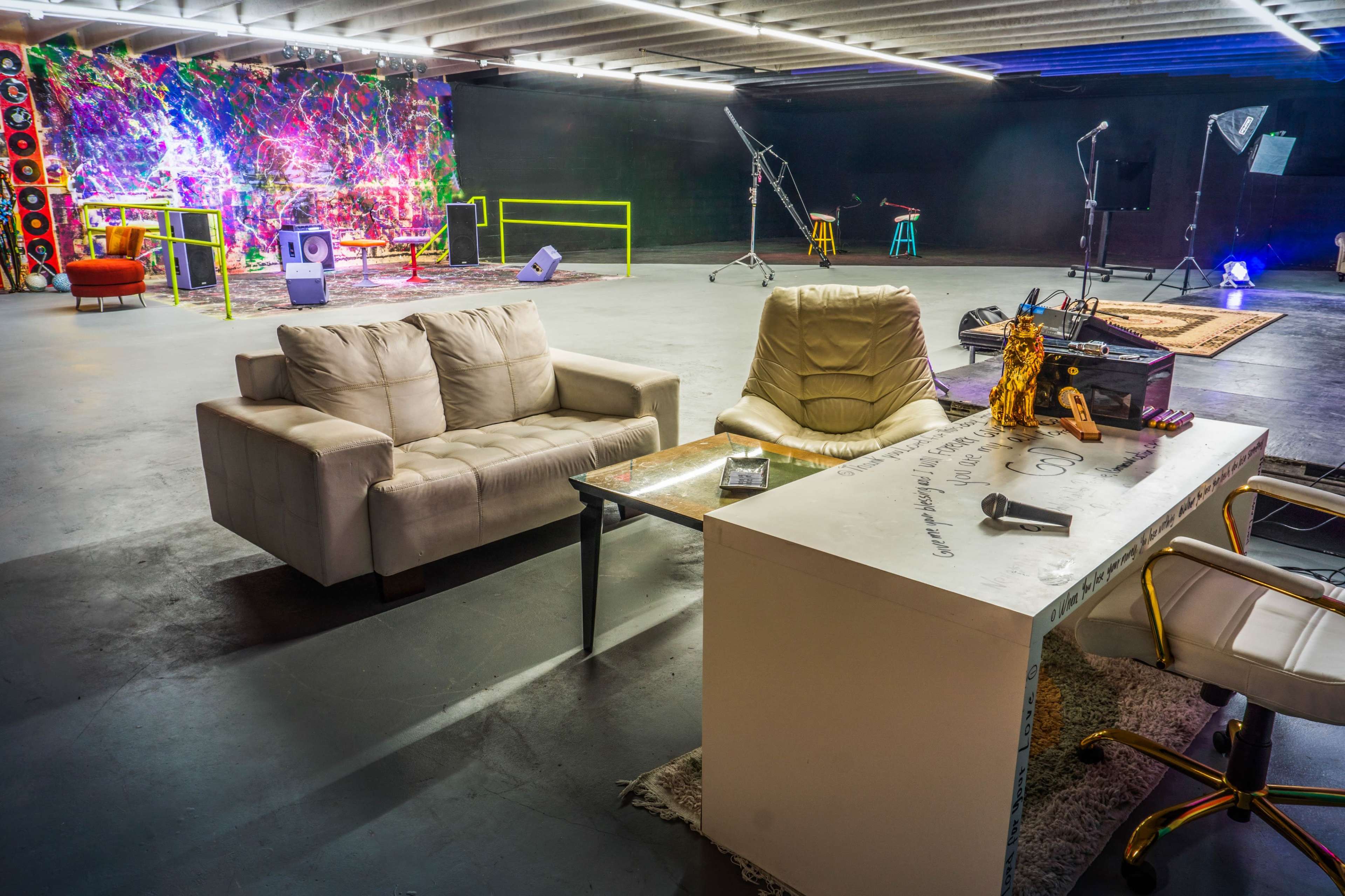 A spacious studio with two sofas, a desk with chairs, and colorful lighting in the background.