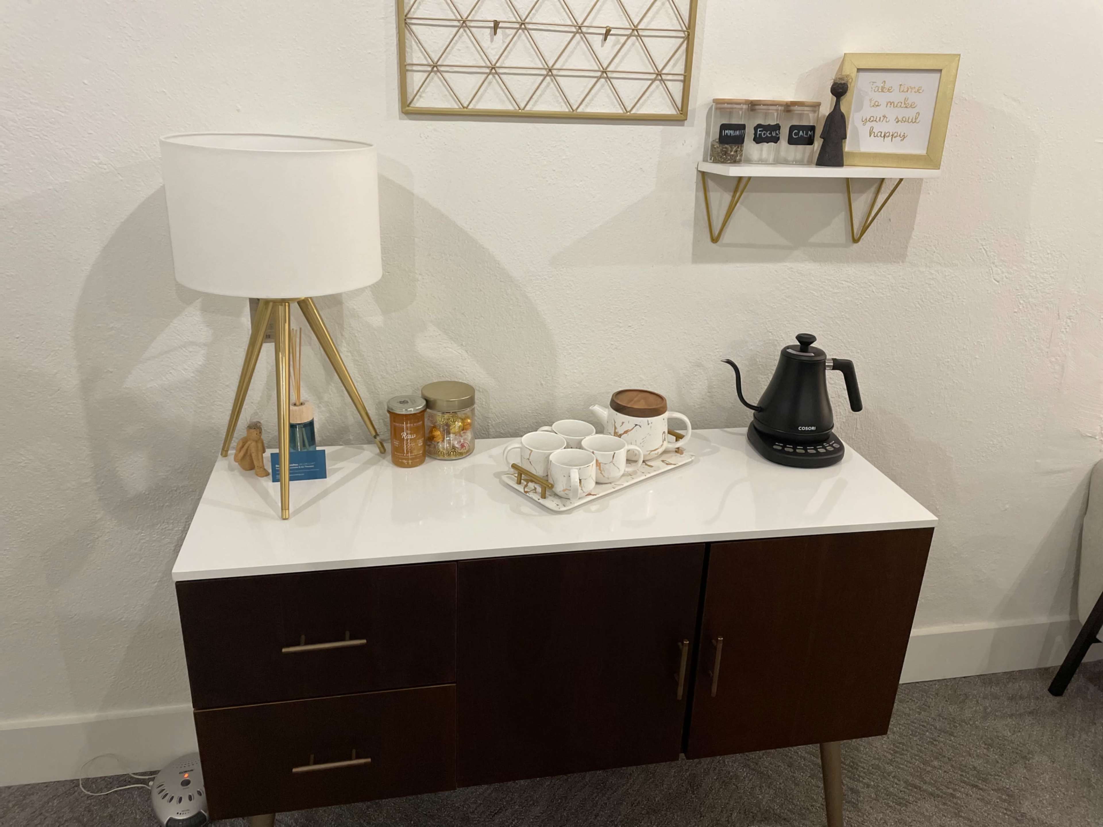 A mid-century modern sideboard with a white tabletop displays a lamp, jars, tea cups, and a kettle against a textured wall.