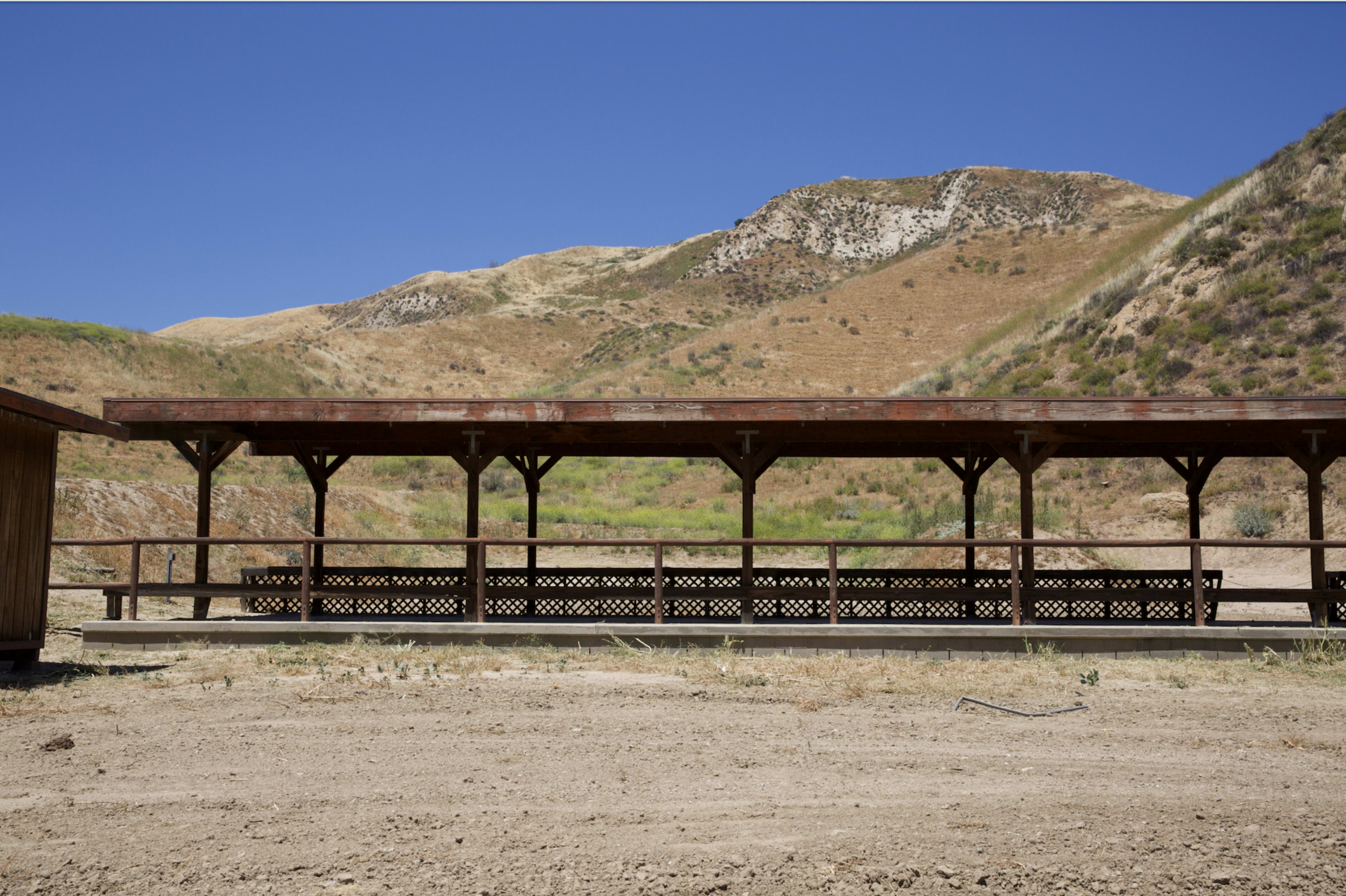 A wooden pavilion with benches is situated in a dry, hilly landscape under a clear blue sky.