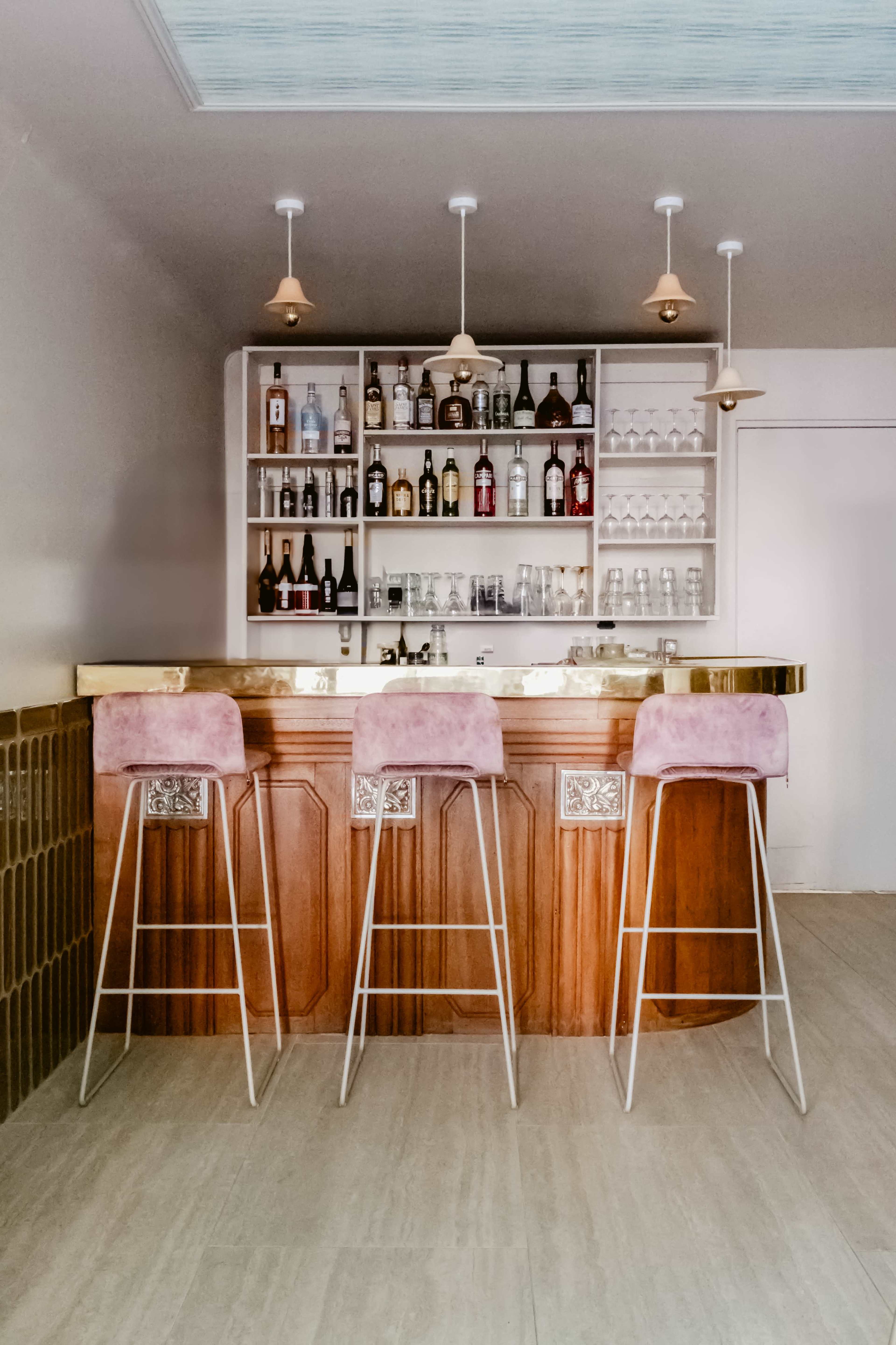 A modern bar features a wooden counter with three pink upholstered stools and a wall of shelves stocked with various liquor bottles and glassware.
