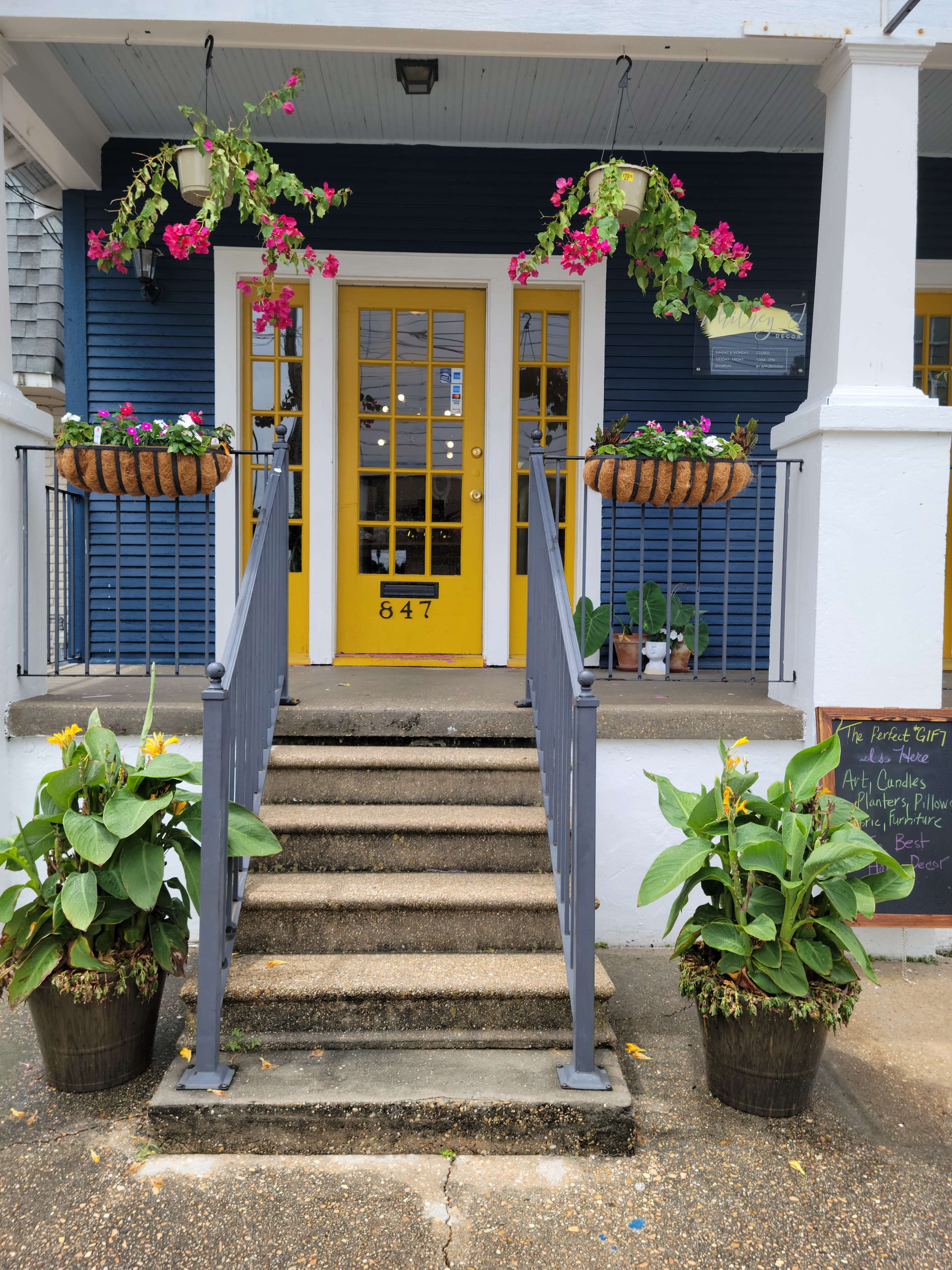 The image shows a set of outdoor steps leading up to a blue building with a yellow door, flanked by hanging flower pots and potted plants at the base.