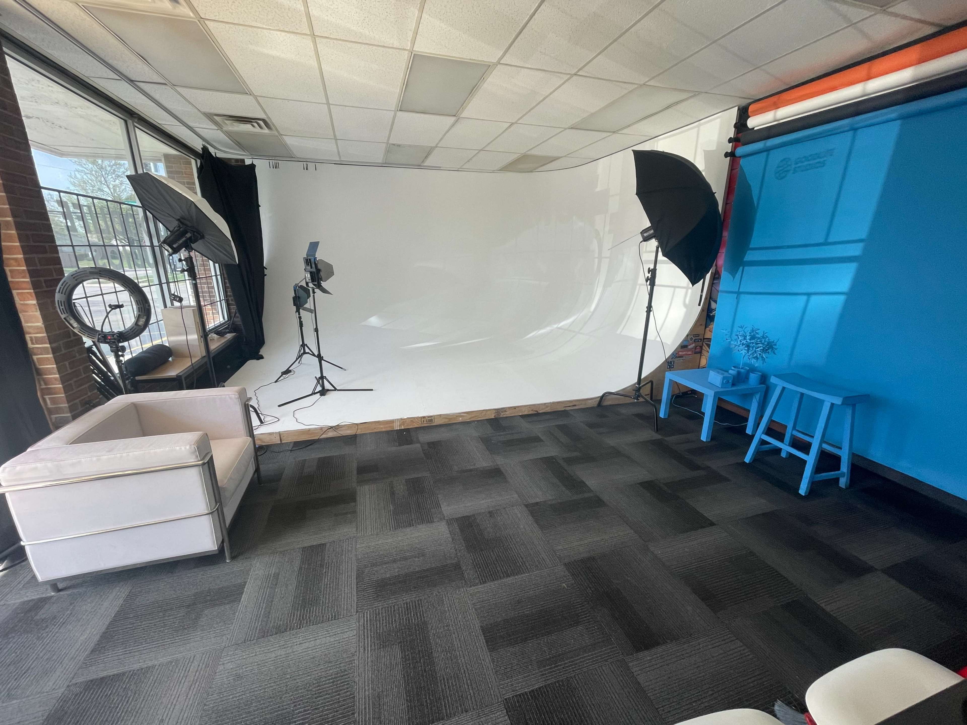 A photography studio with a white backdrop, softbox lights, and a blue wall, featuring a white sofa and two blue chairs.