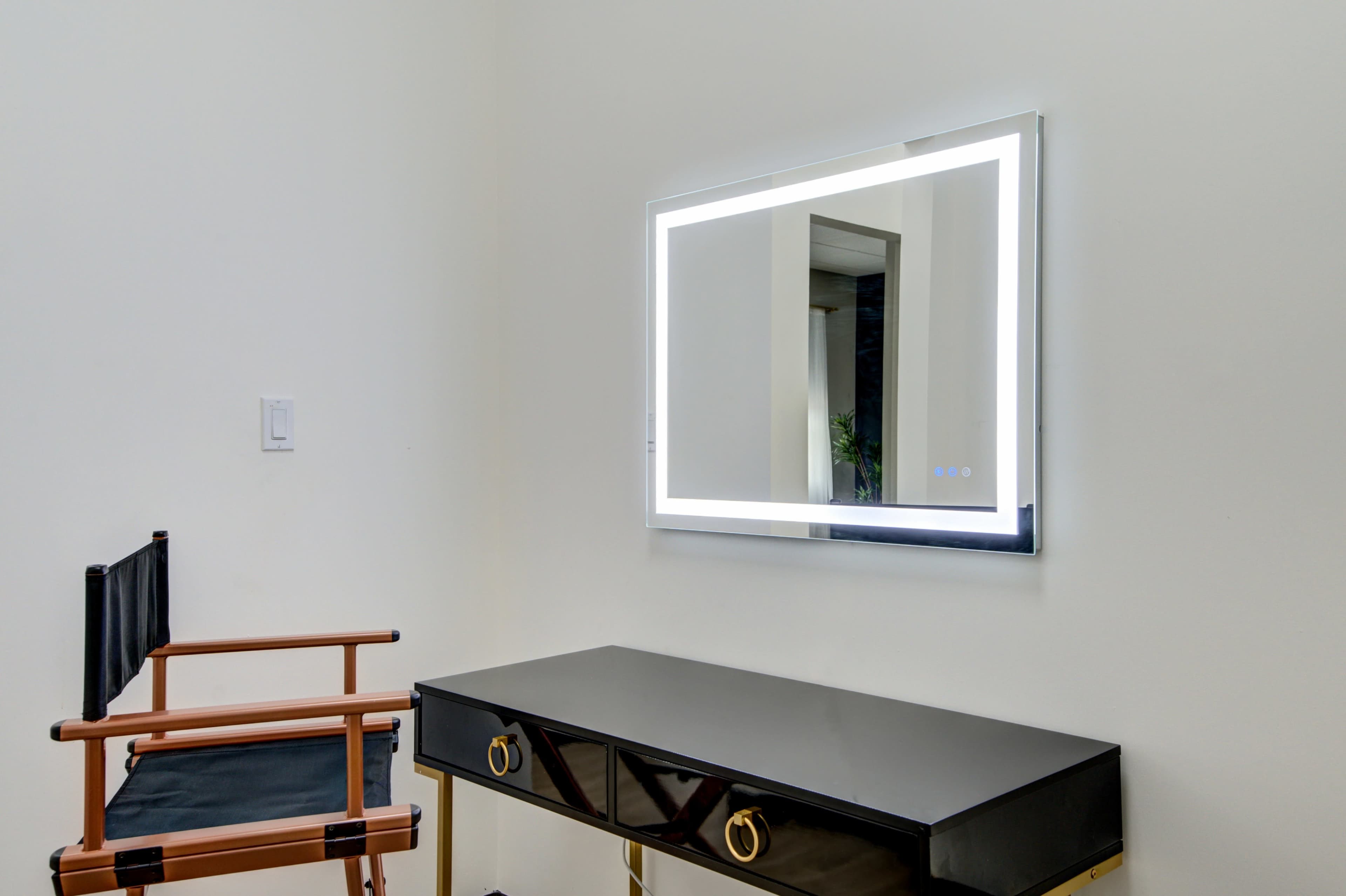 A modern vanity table with a mirror and an illuminated frame is positioned next to a wooden director's chair against a plain wall.
