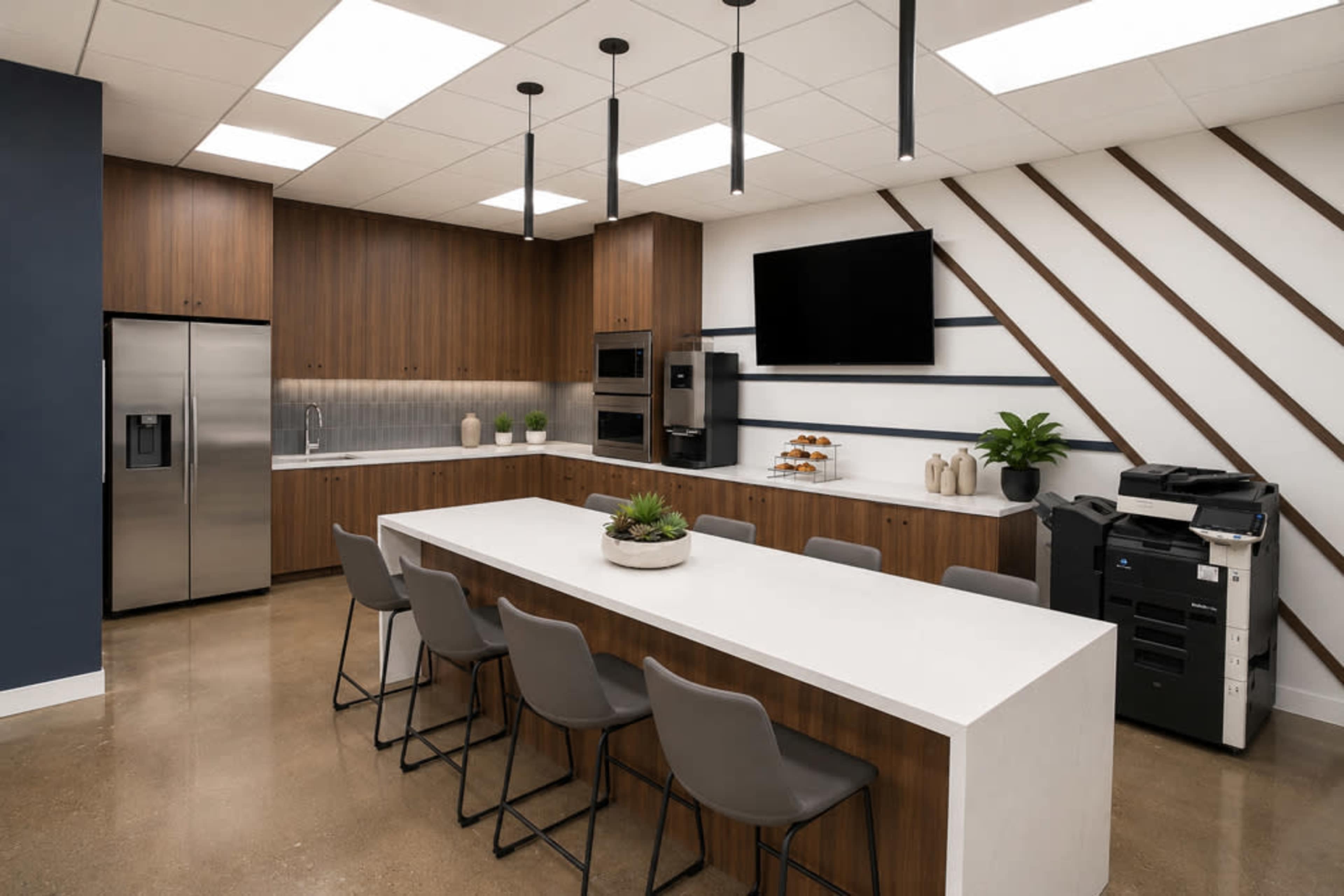 The image shows a modern kitchen with wooden cabinetry, a large white countertop table, seating for eight, and appliances including a refrigerator and oven, alongside a printer and decor plants.
