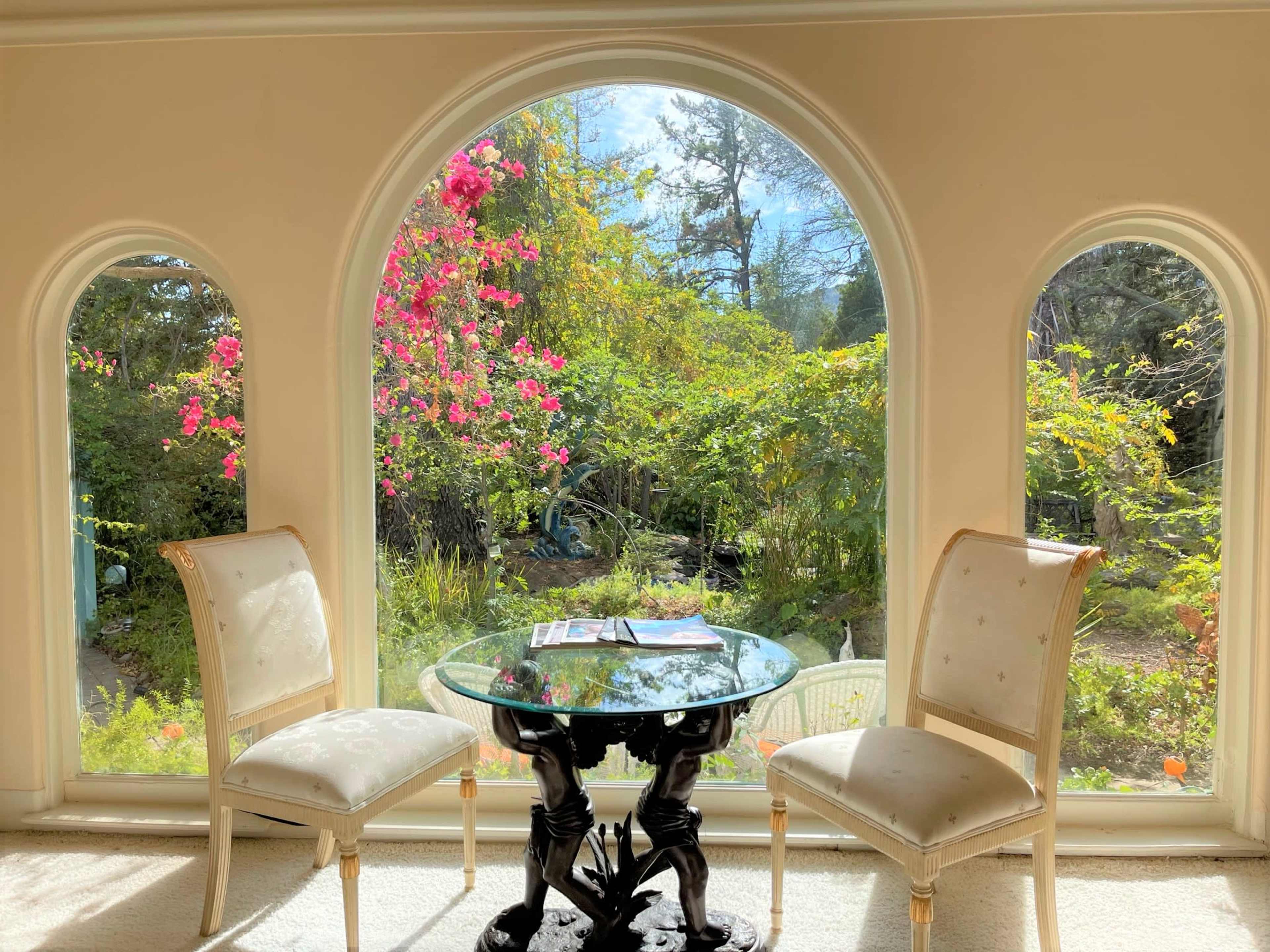A glass table sits between two chairs in front of an arched window, showcasing a garden filled with vibrant flowers and greenery.