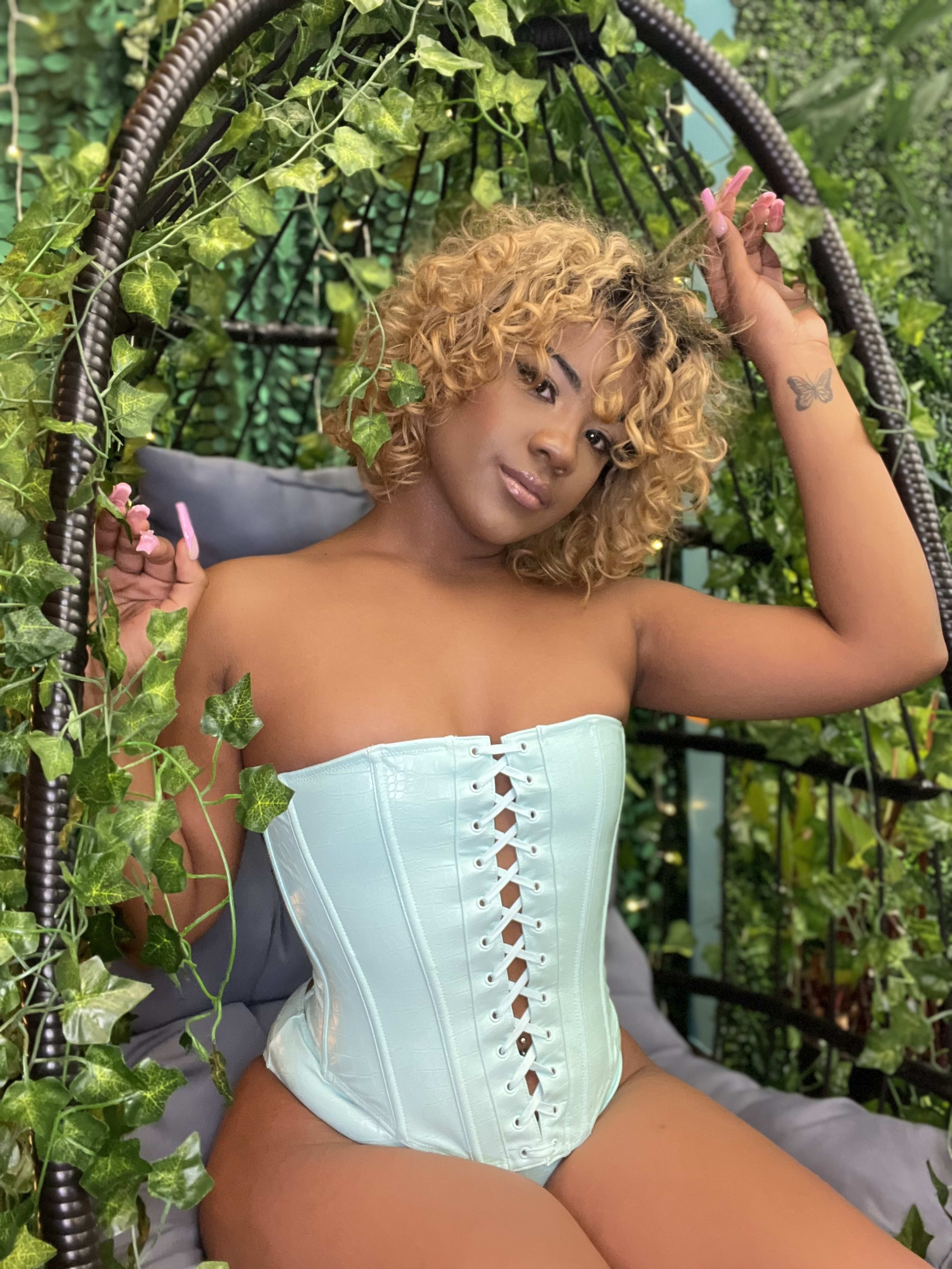 A fashion model in a light blue corset posing outdoors amidst plants.