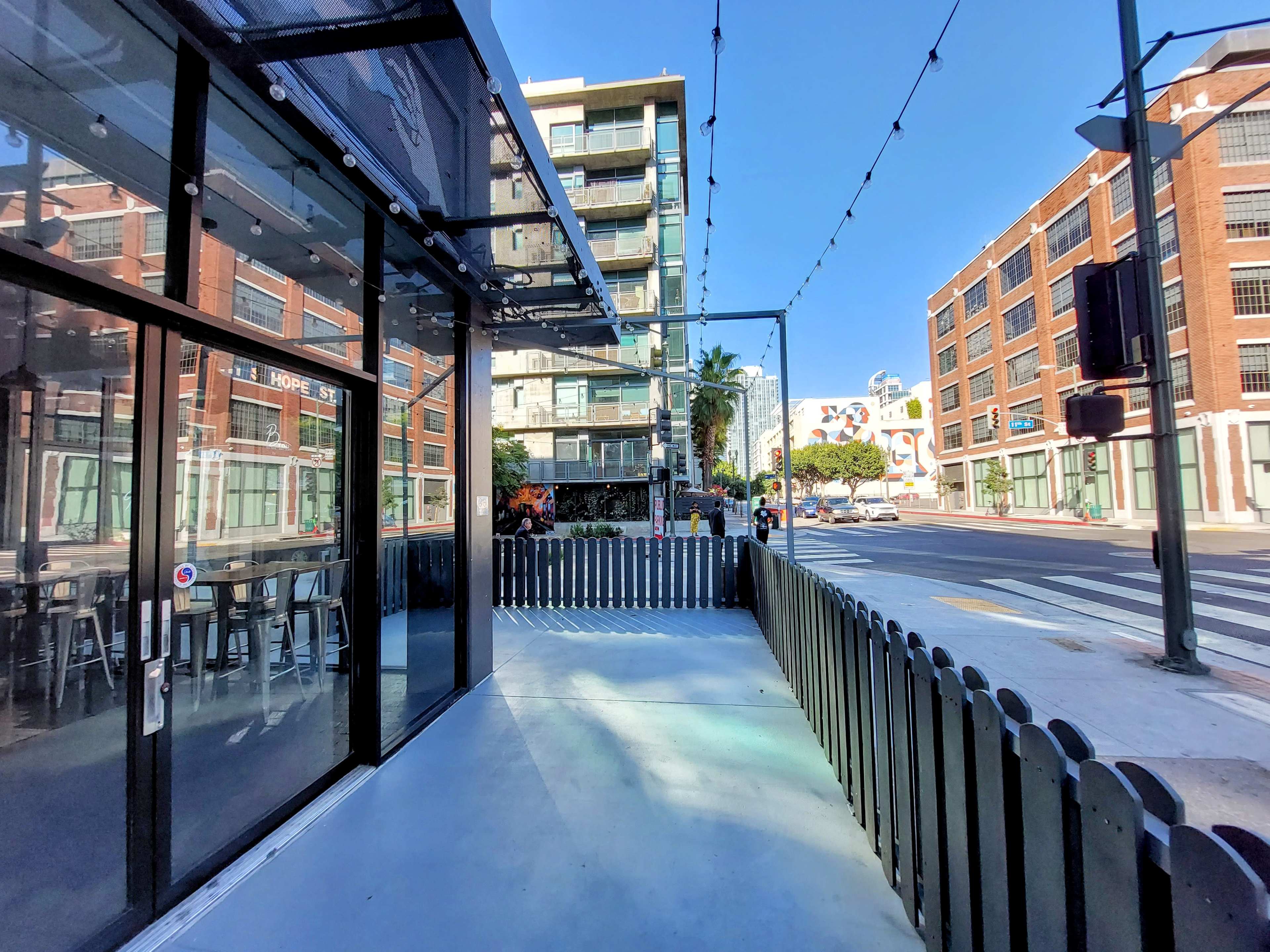A modern outdoor seating area with a glass enclosure facing a street lined with buildings and palm trees.