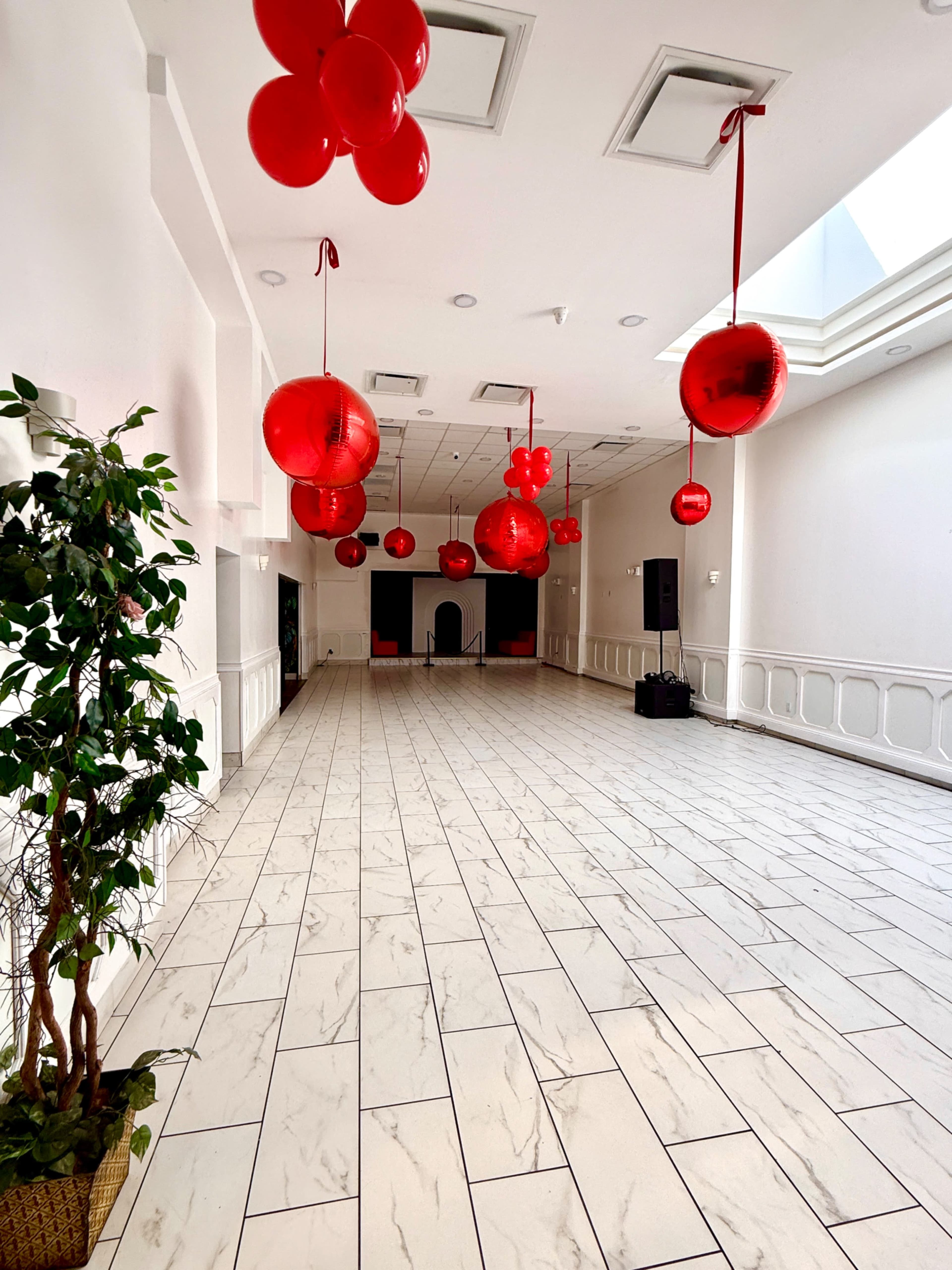 An empty event space features white walls and a tiled floor, adorned with large red balloons hanging from the ceiling.