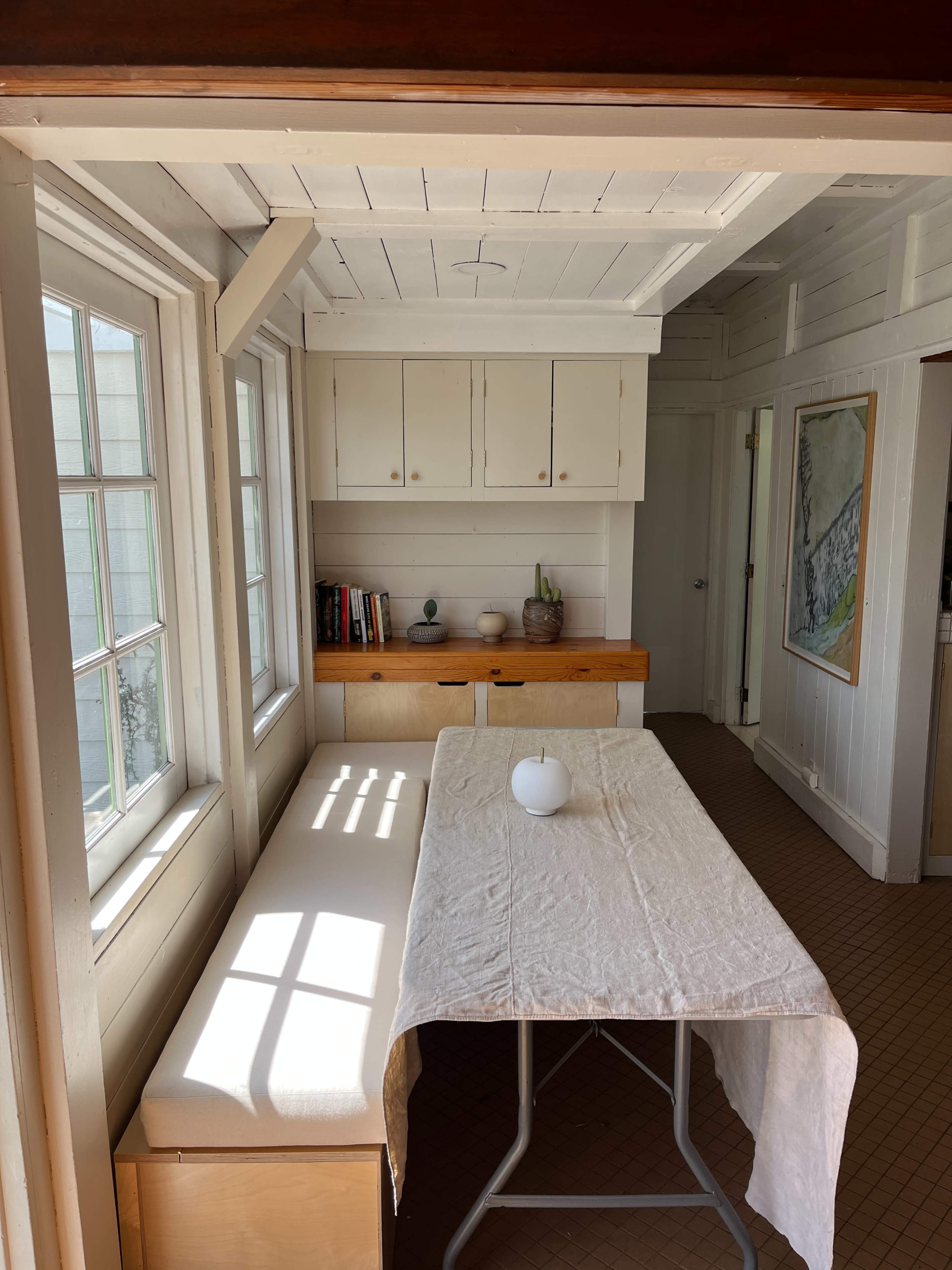 A bright, minimalist kitchen area with a table covered by a white cloth and a bench against the wall, illuminated by natural light through large windows.