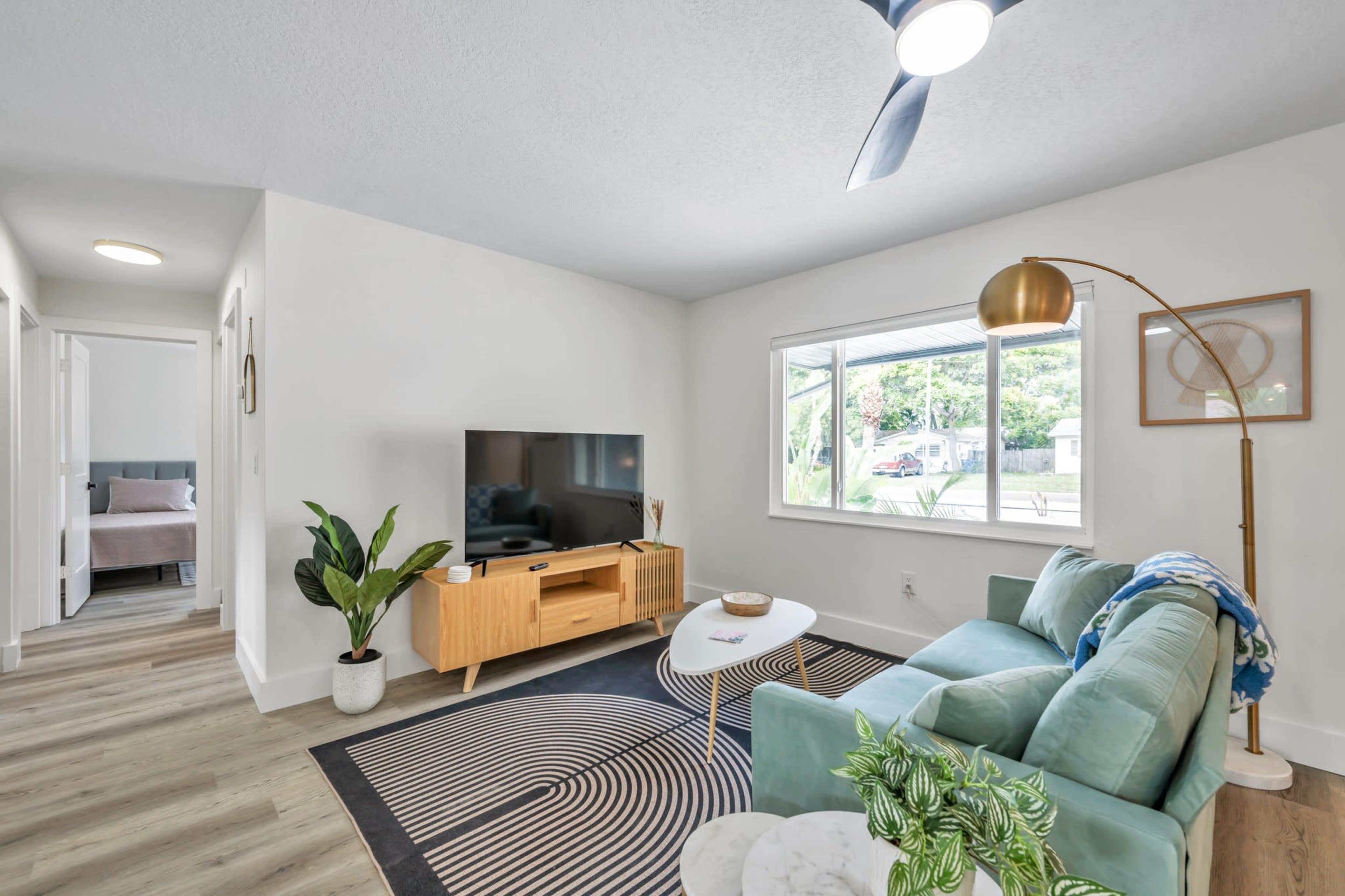 A modern living room with a teal sofa, a wooden TV stand, a round coffee table, and a large window providing natural light.