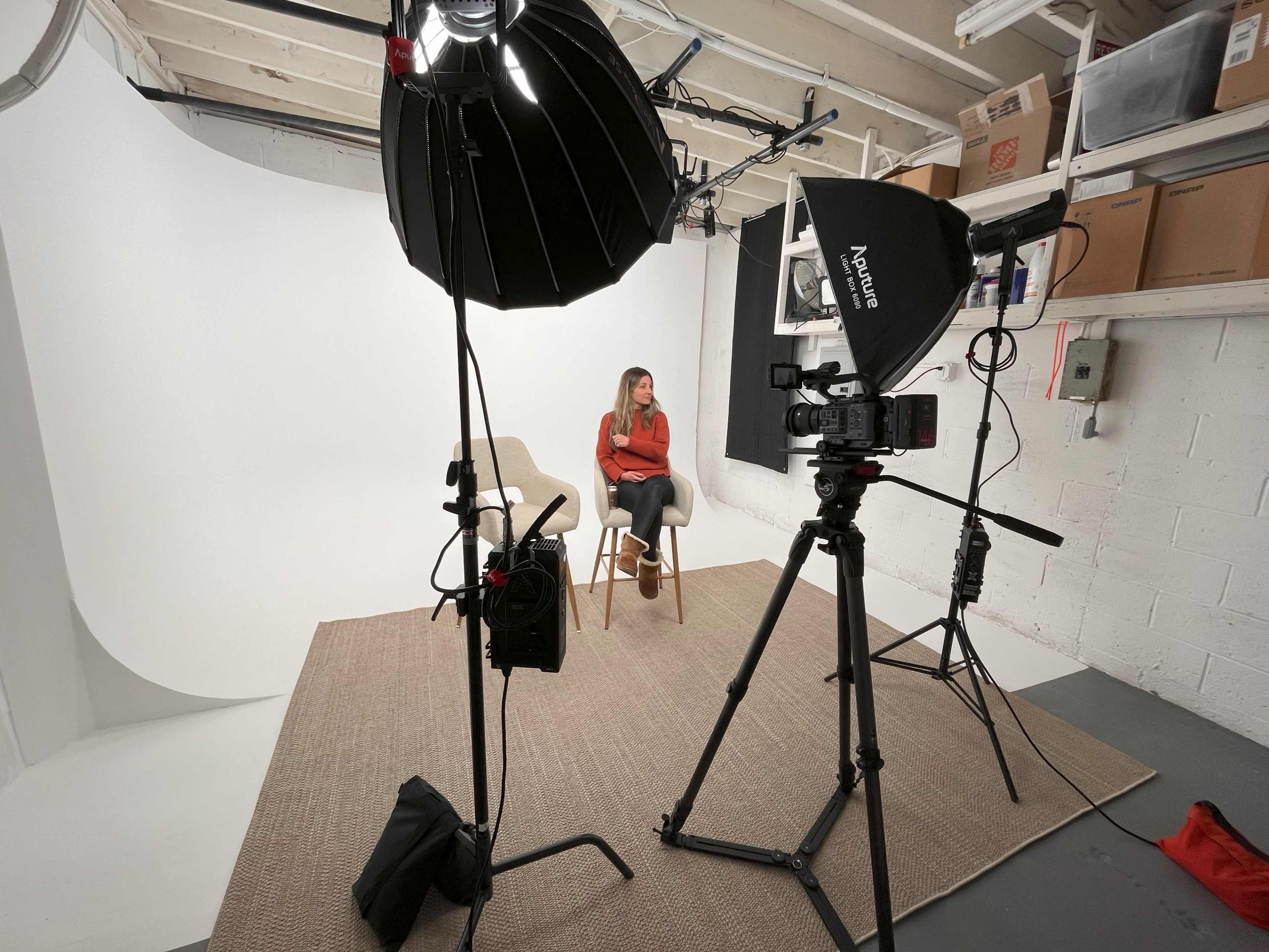 A woman sits on a chair in a photography studio equipped with lighting and a backdrop.