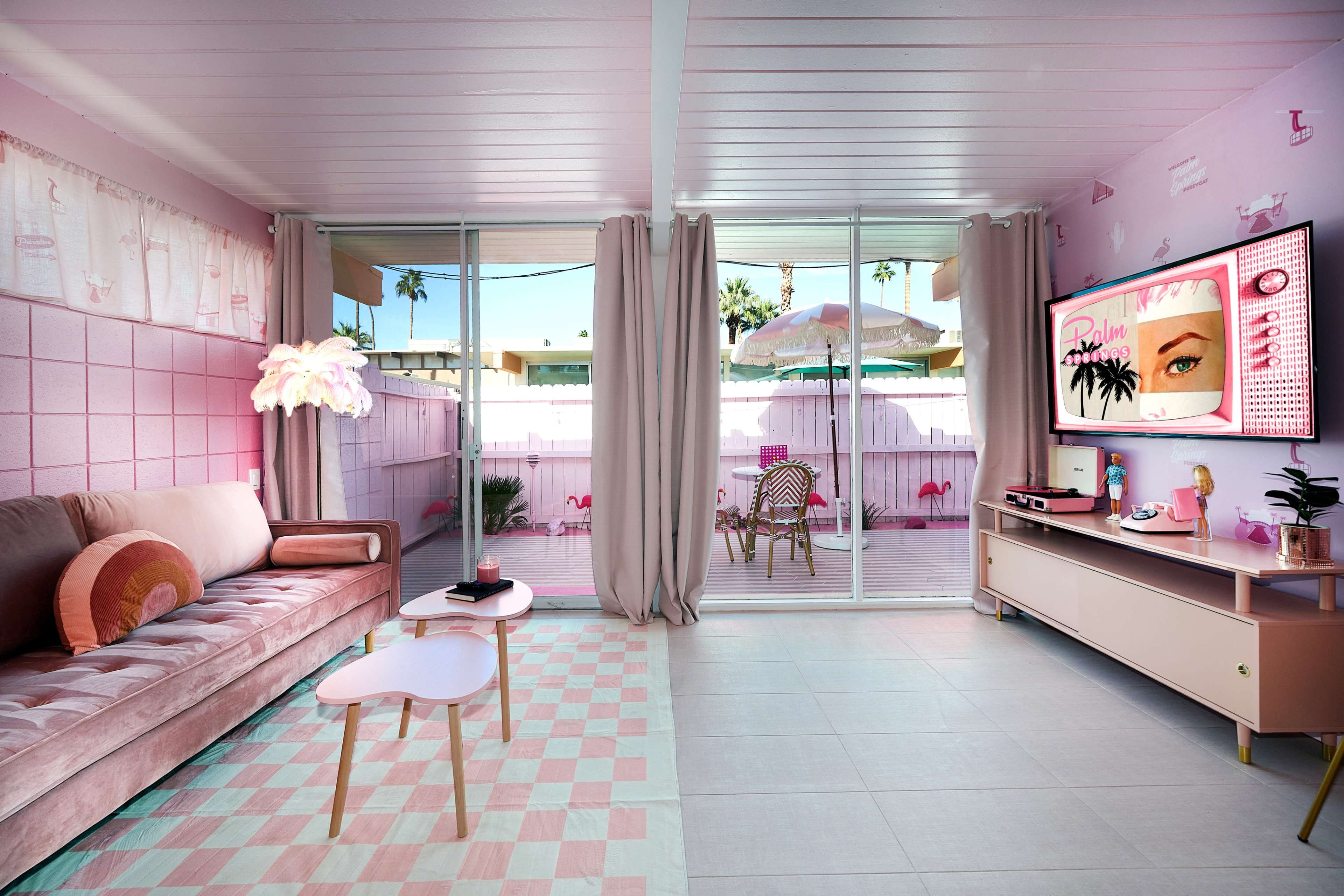 A brightly colored living room features a pink sofa, a small coffee table, and large windows that open to an outdoor patio with flamingo decorations.