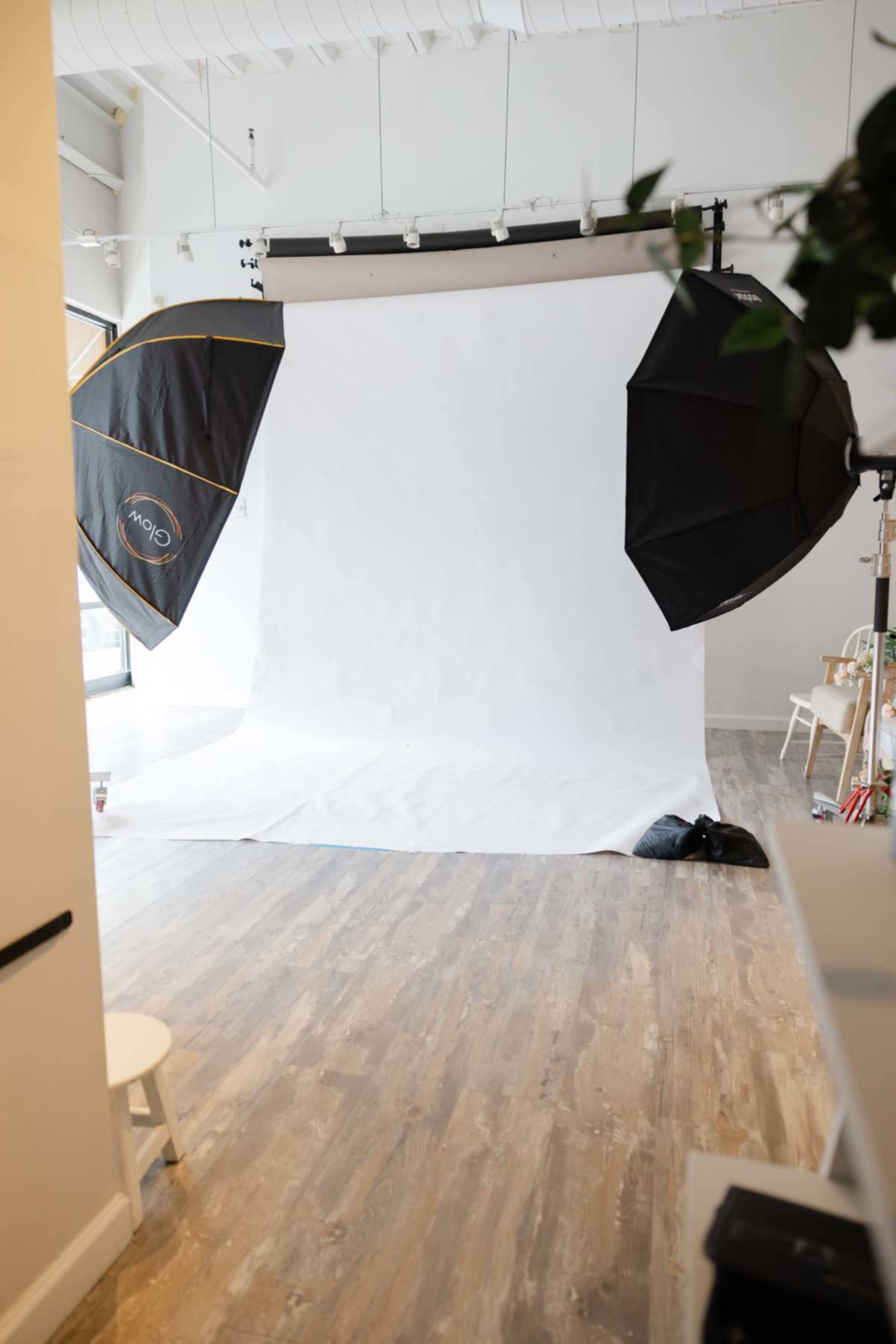 The image shows a photography studio with a white backdrop, two softbox lights positioned on either side, and wooden flooring.