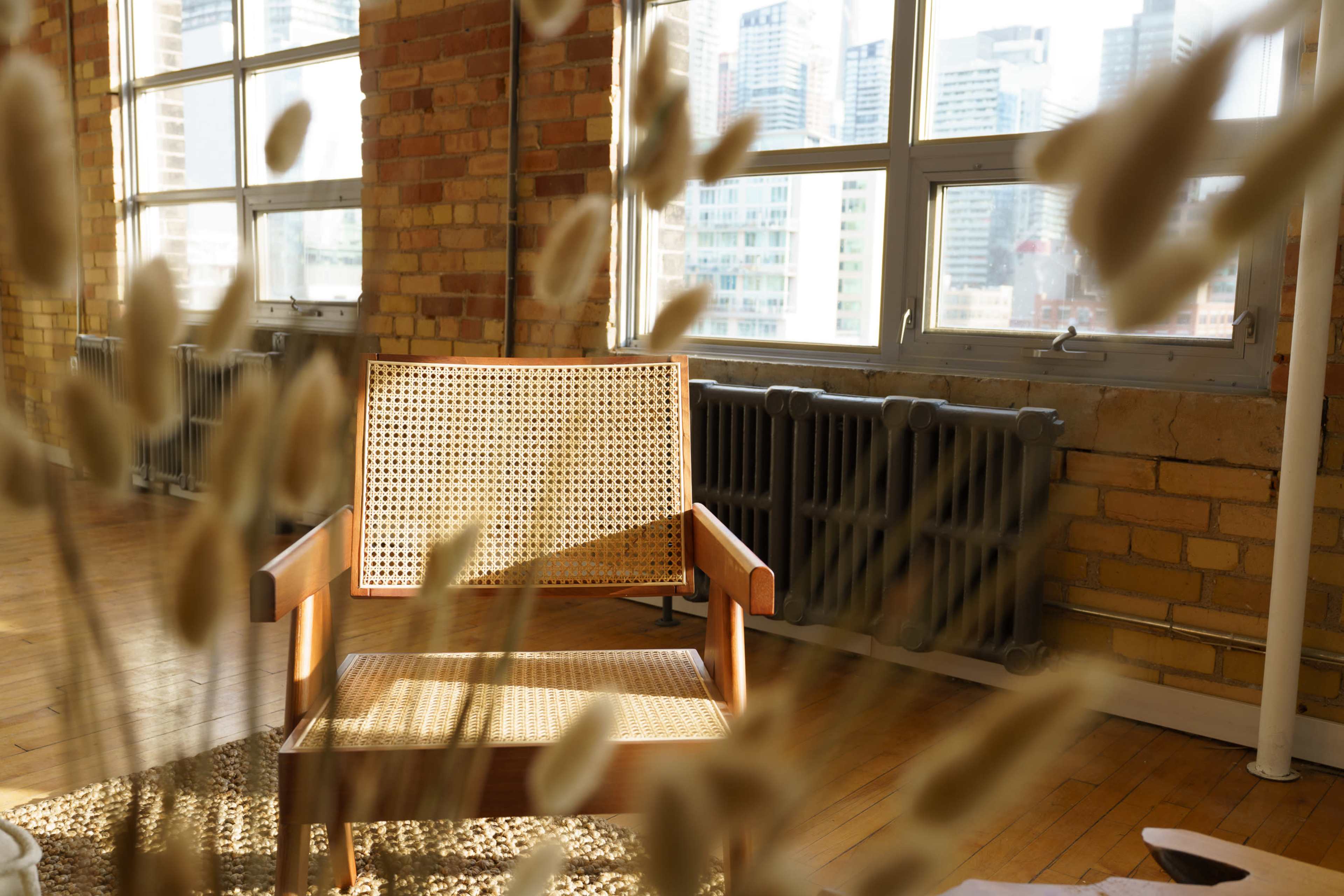 A wooden chair with a woven backrest is positioned in a bright room filled with large windows overlooking a cityscape.