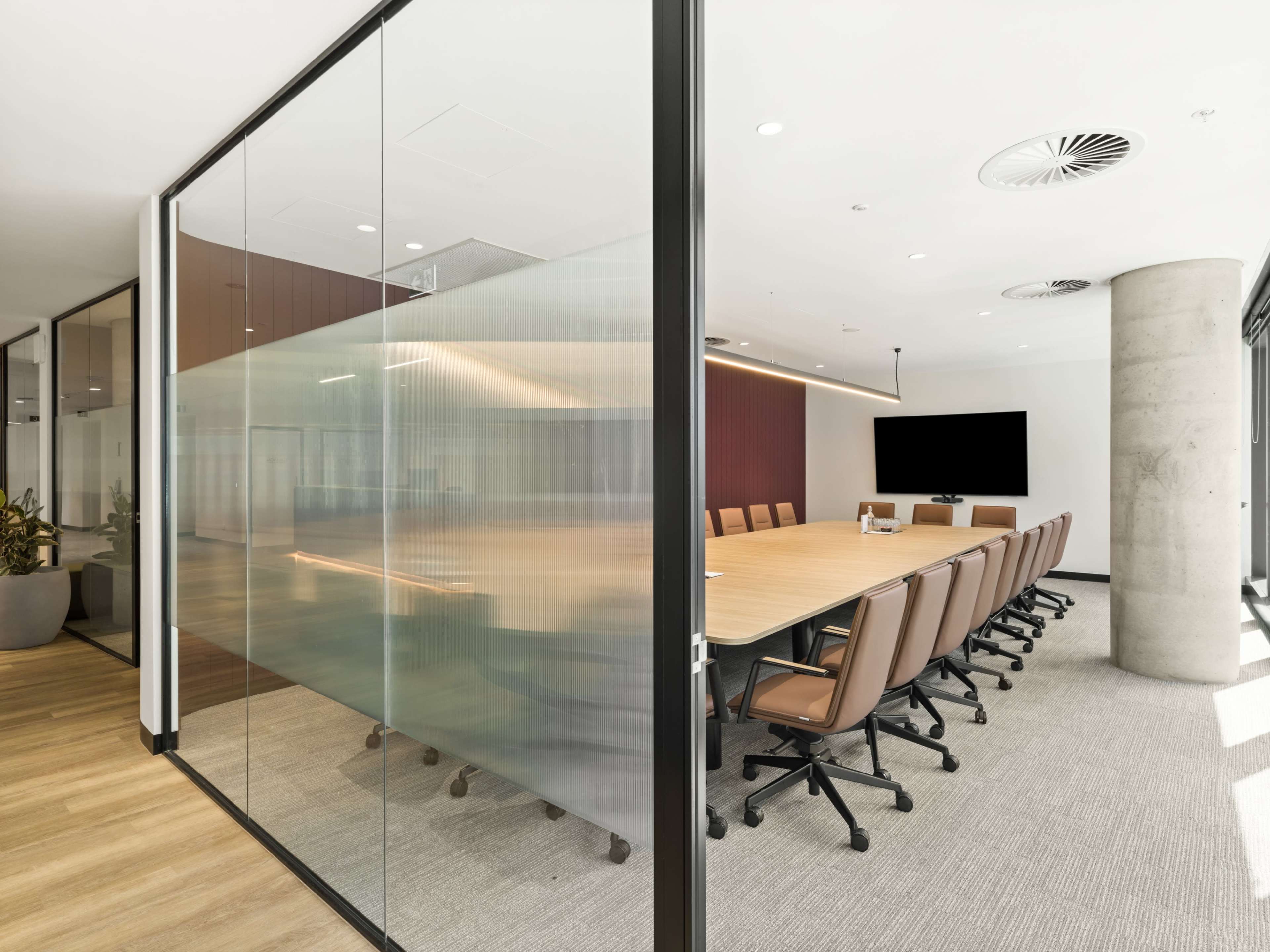 A modern conference room features a large wooden table surrounded by leather chairs, enclosed by glass walls and illuminated by ceiling lights.