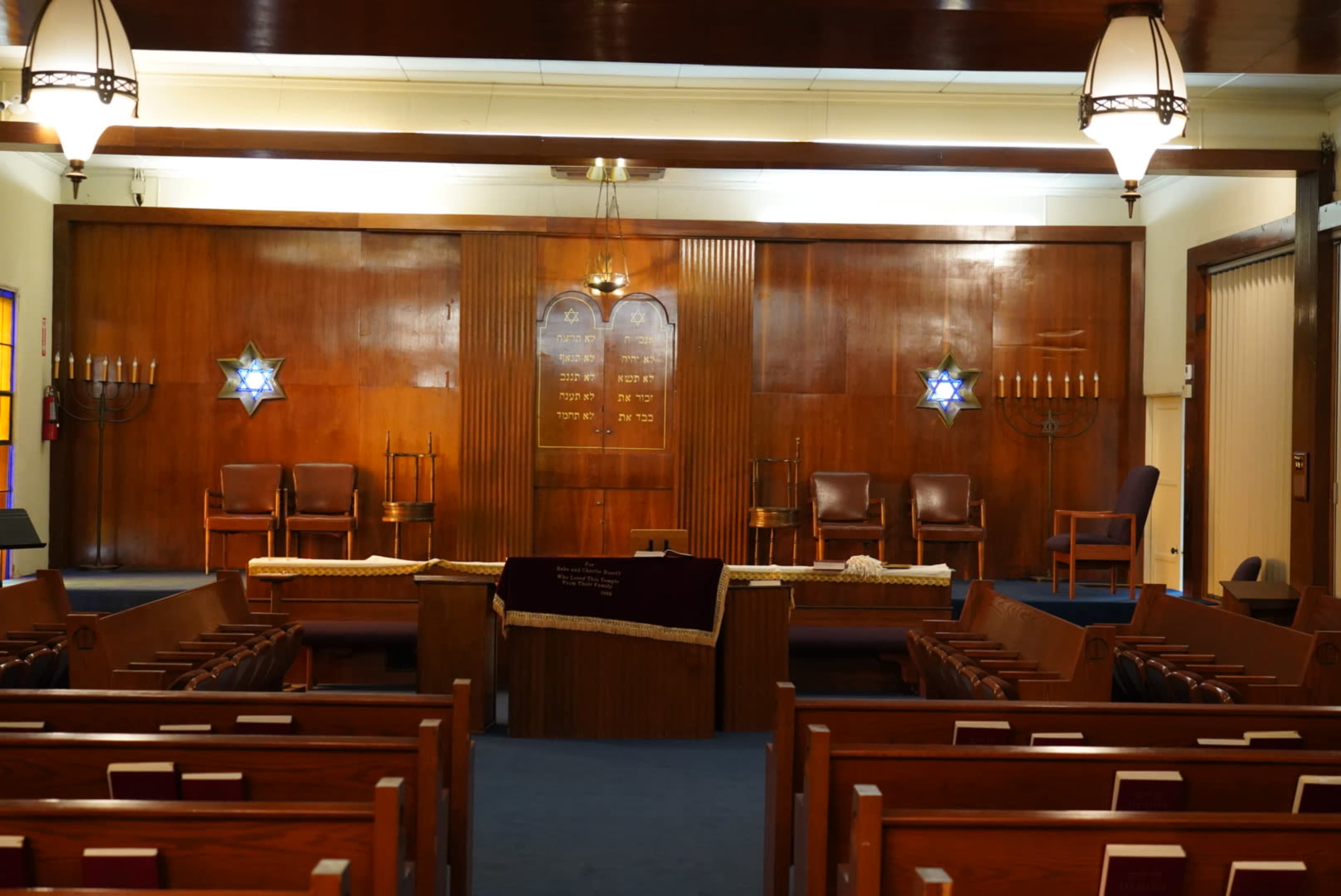 The interior of a synagogue features wooden paneling, chairs, a central table, and decorative stars of David on the walls.