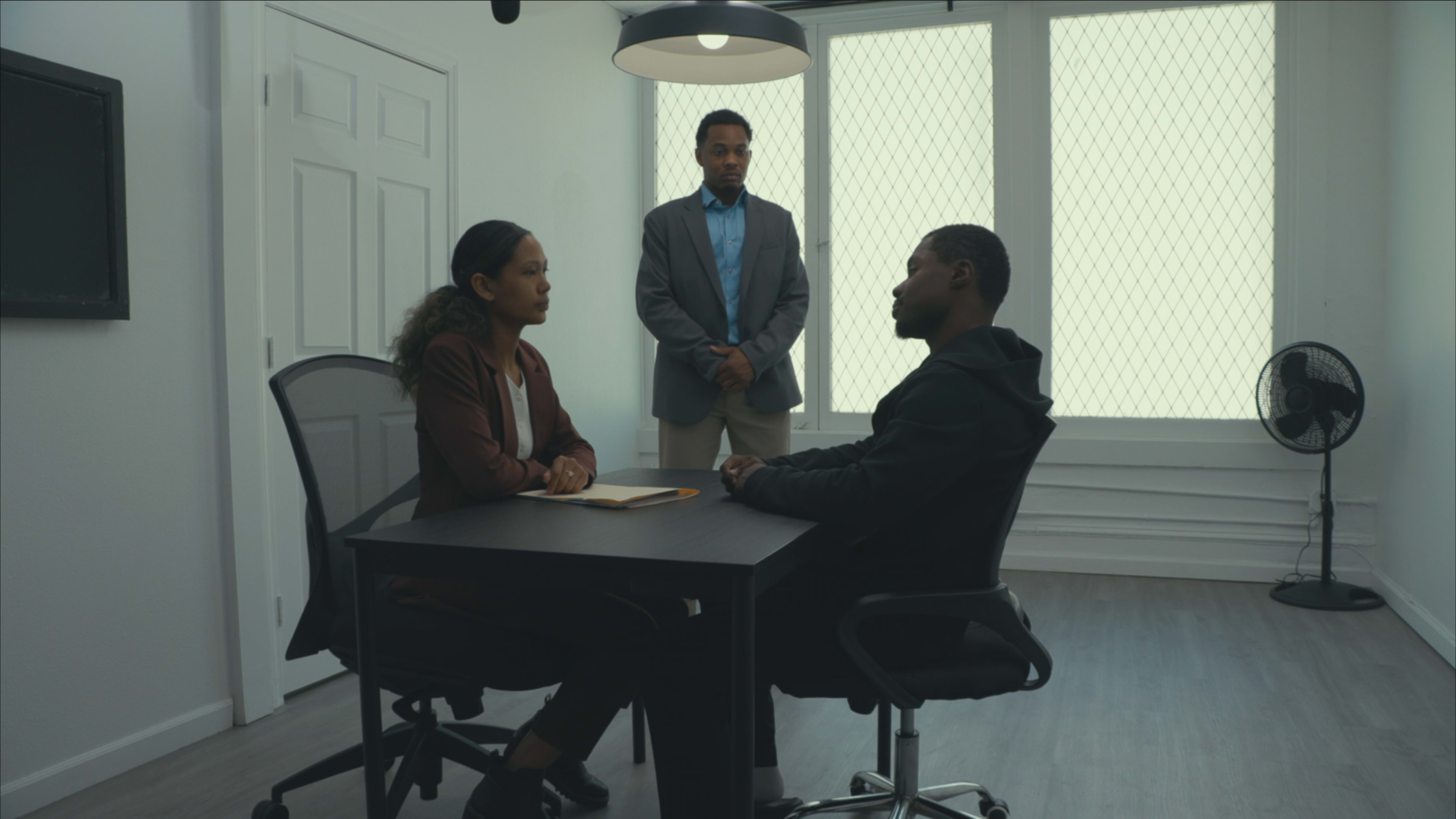 A woman in business attire sits at a table across from a man in casual clothing, while a man in a suit stands behind them in a well-lit office.