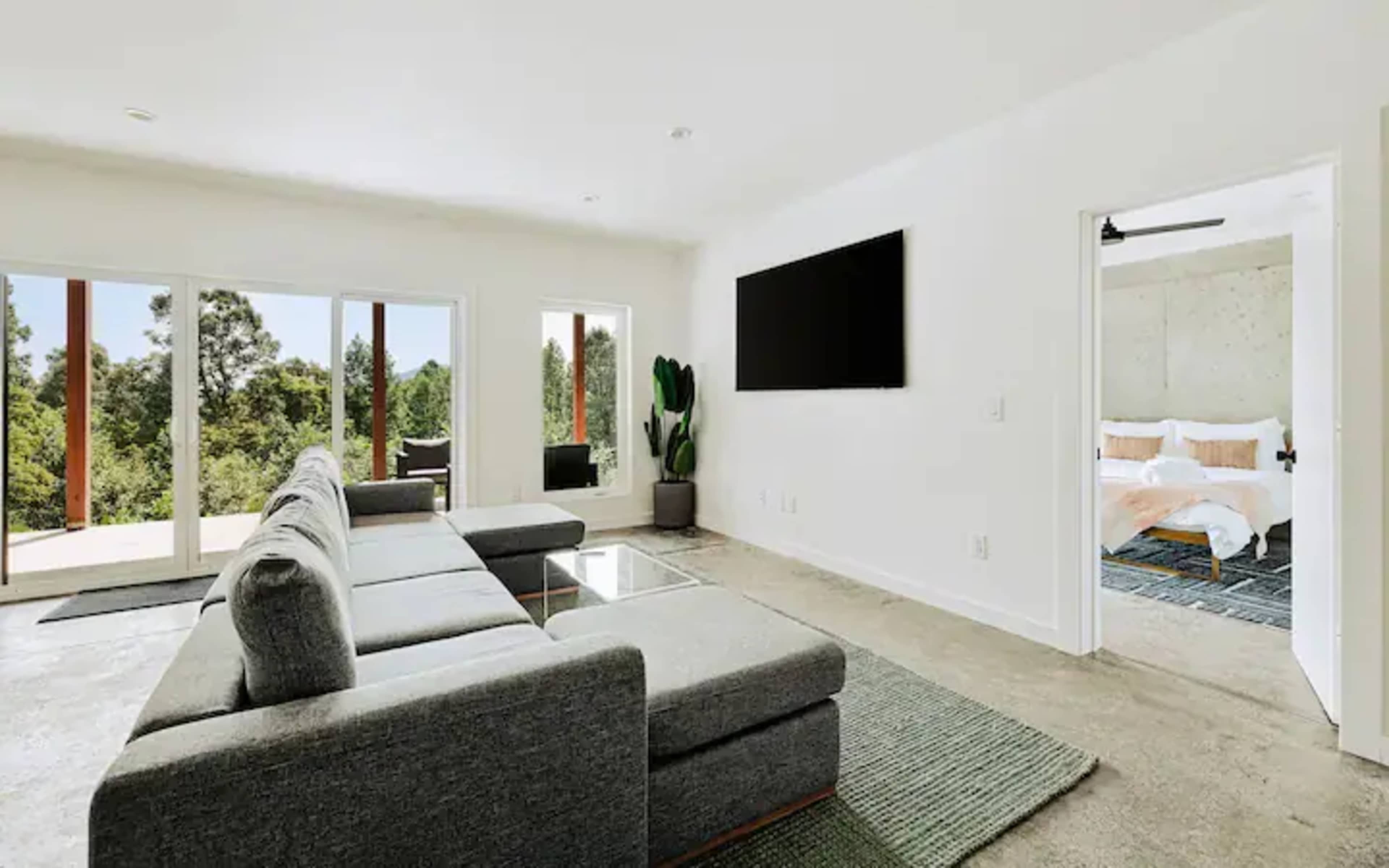 A modern living room features a gray sectional sofa, a coffee table, a large flat-screen TV on the wall, and doors leading to an outdoor view.