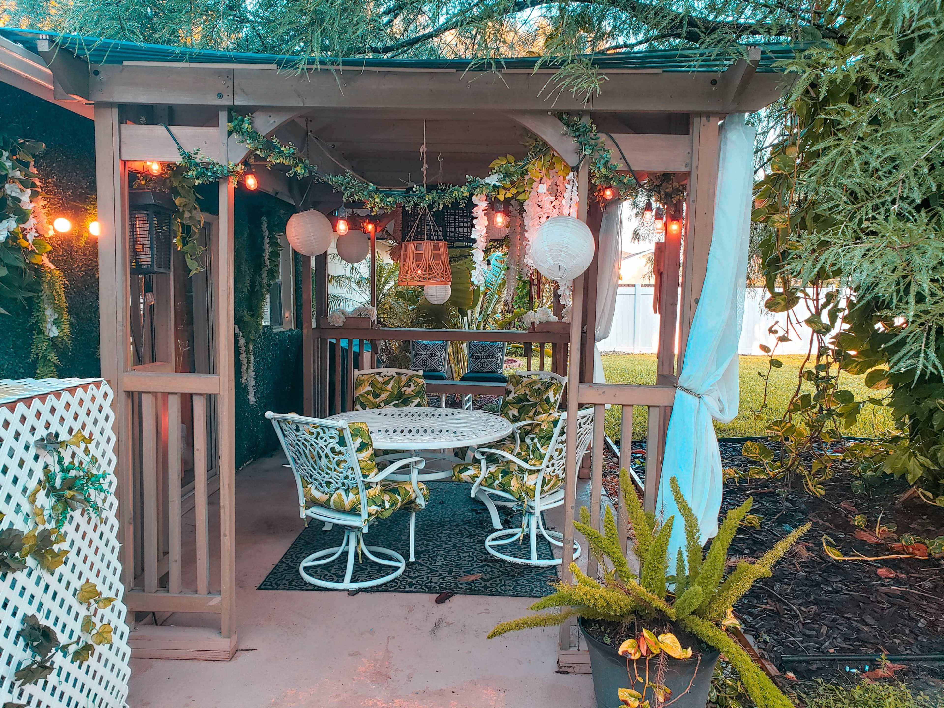 A cozy outdoor seating area features a round table surrounded by four chairs, adorned with green and white cushions, under string lights and decorative lanterns.