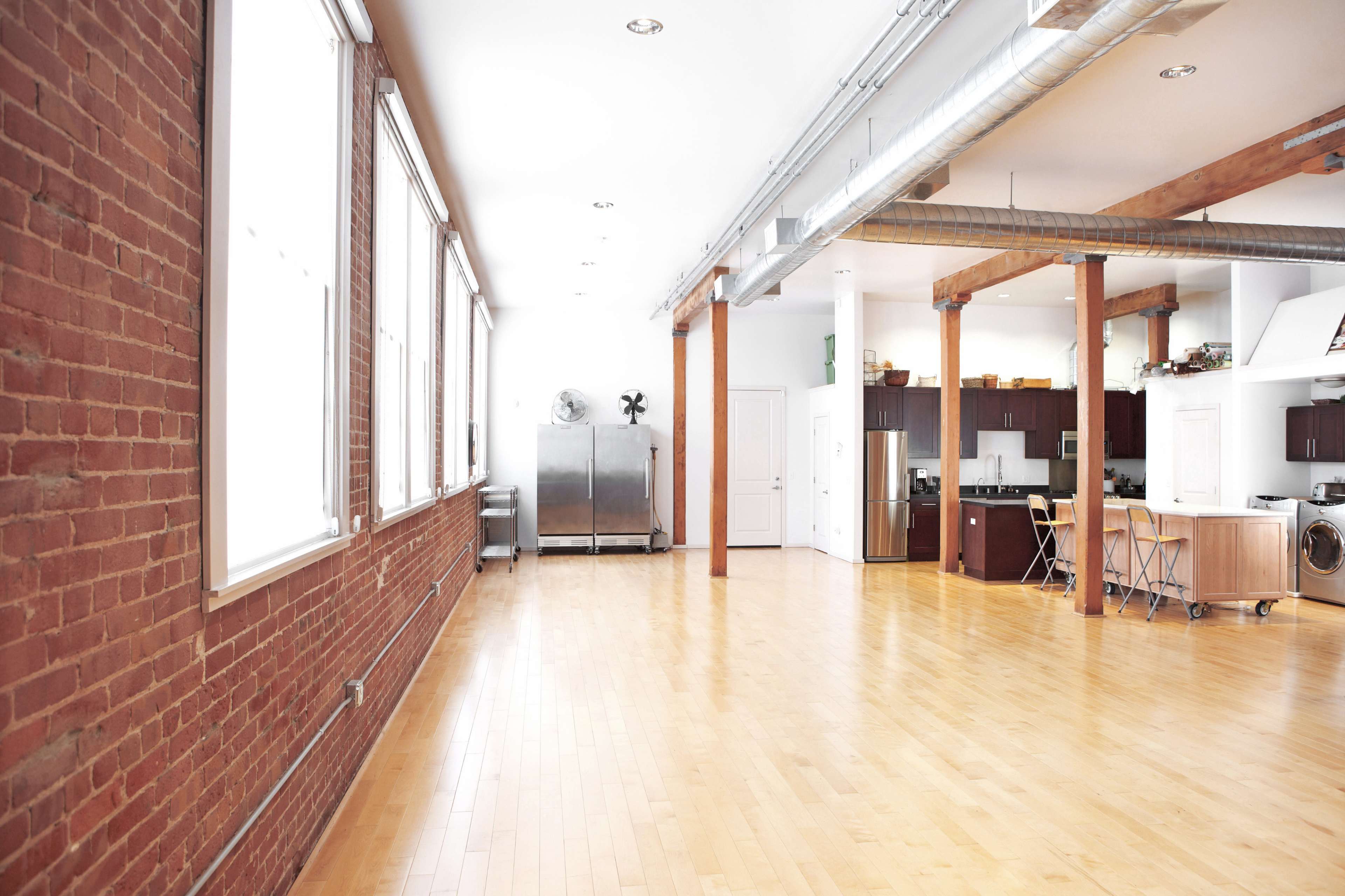 A spacious, modern interior with hardwood floors, large windows, exposed brick walls, and a kitchenette area.