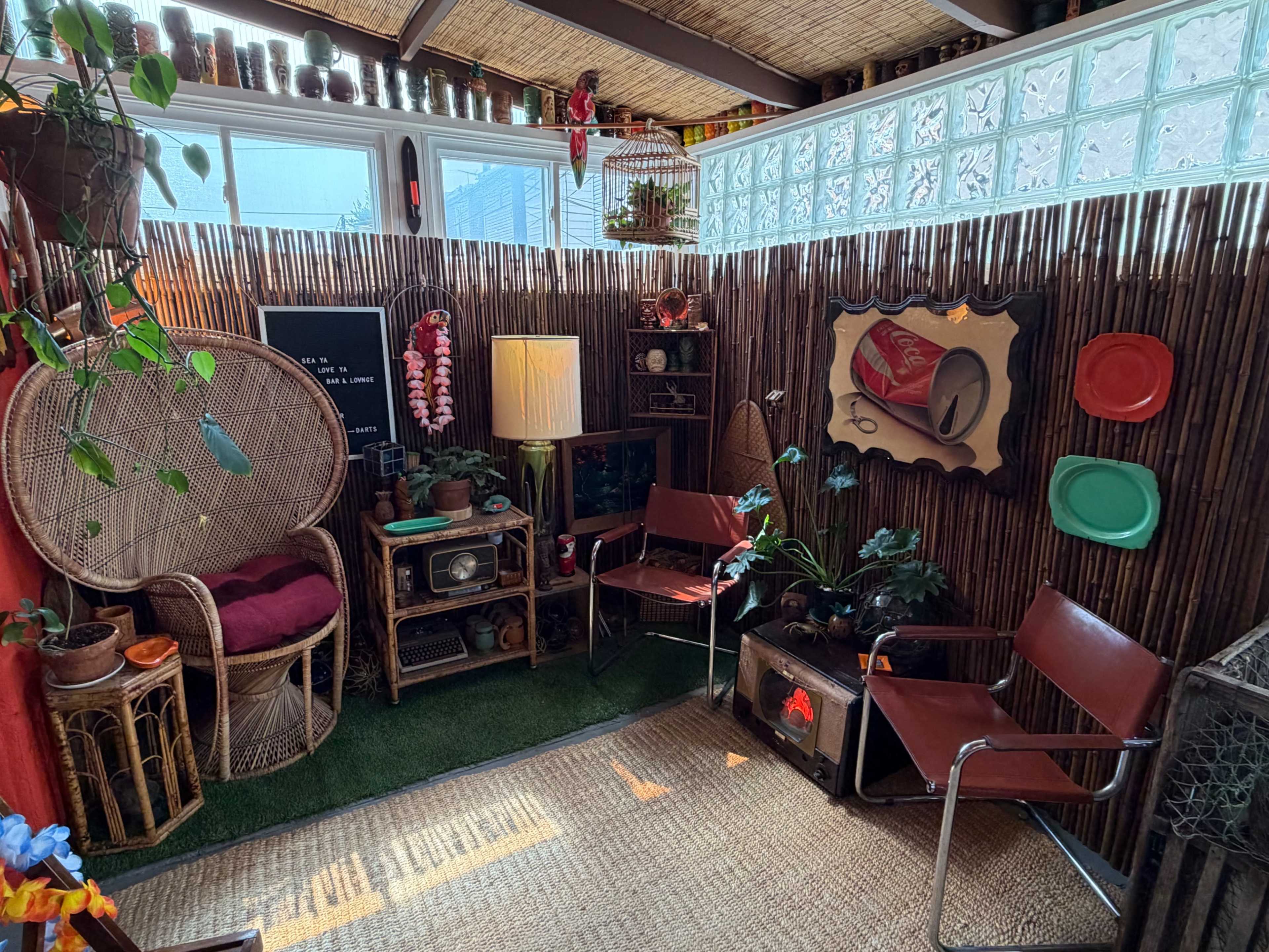 The image shows a cozy indoor space featuring vintage decor, plants, and a seating arrangement with wicker and metal chairs.