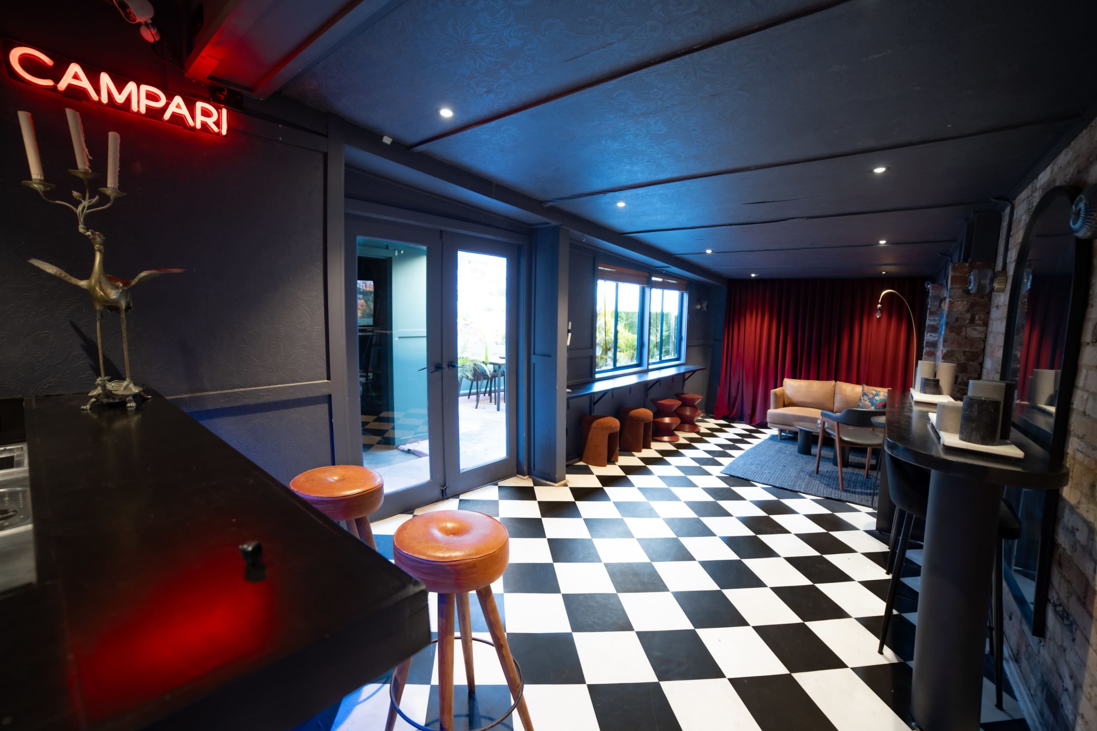 The image shows a modern interior space with a checkered black and white floor, a bar area featuring bar stools, and a seating area with a couch and red curtains.