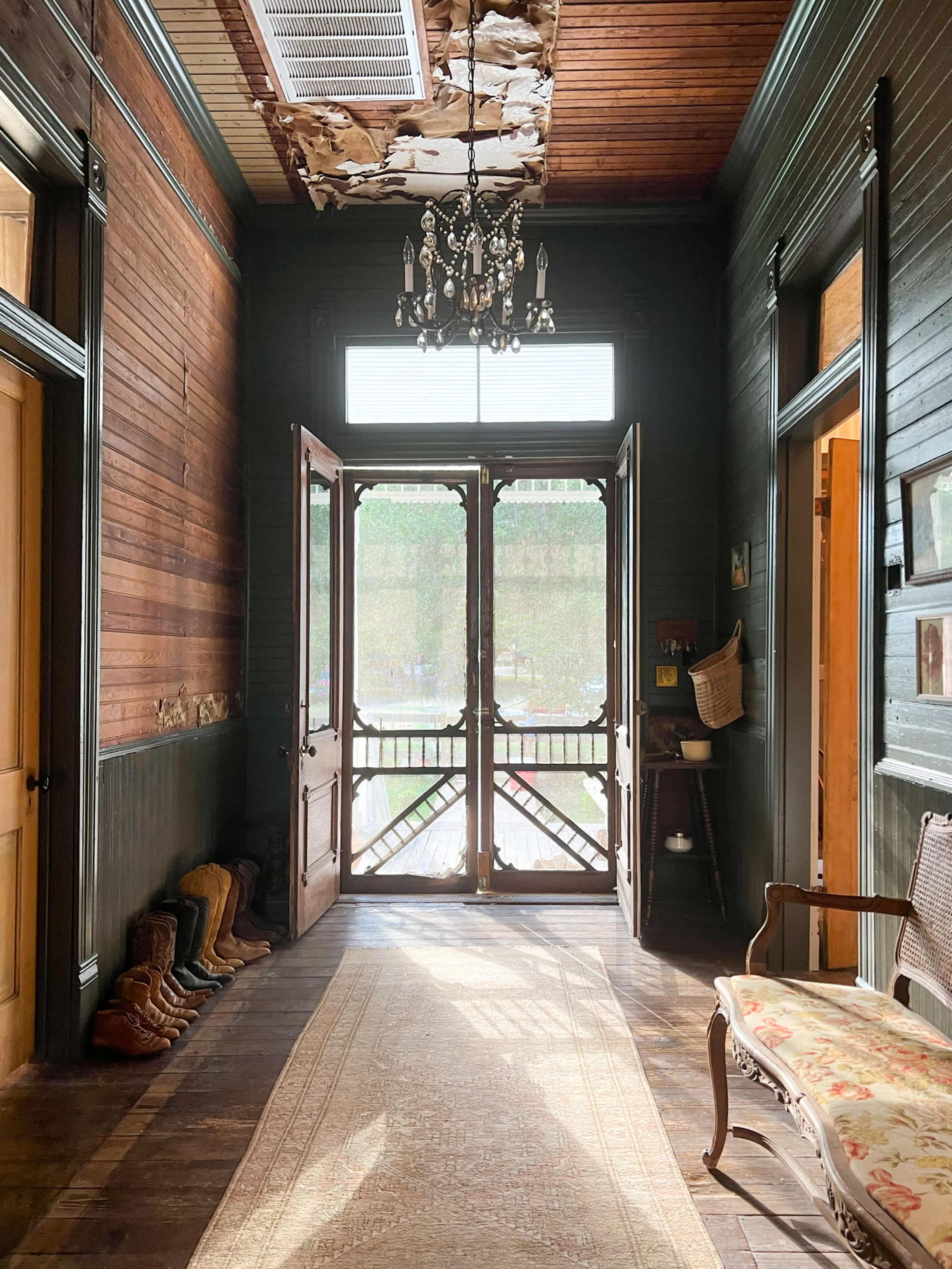 A hallway with wooden walls and a chandelier, featuring two open doors leading outside and a patterned rug on the floor.