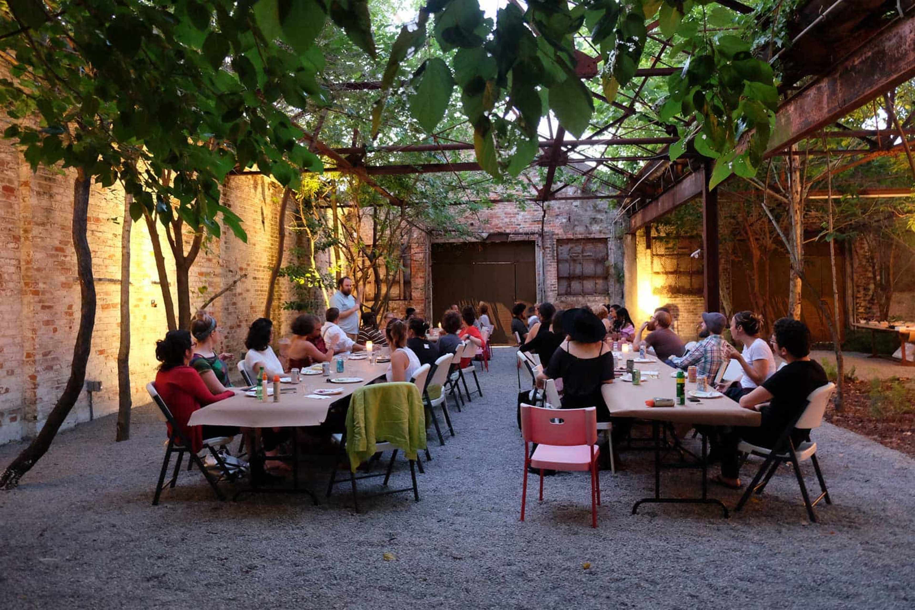 A group of people is seated around tables in an outdoor space surrounded by brick walls and greenery.