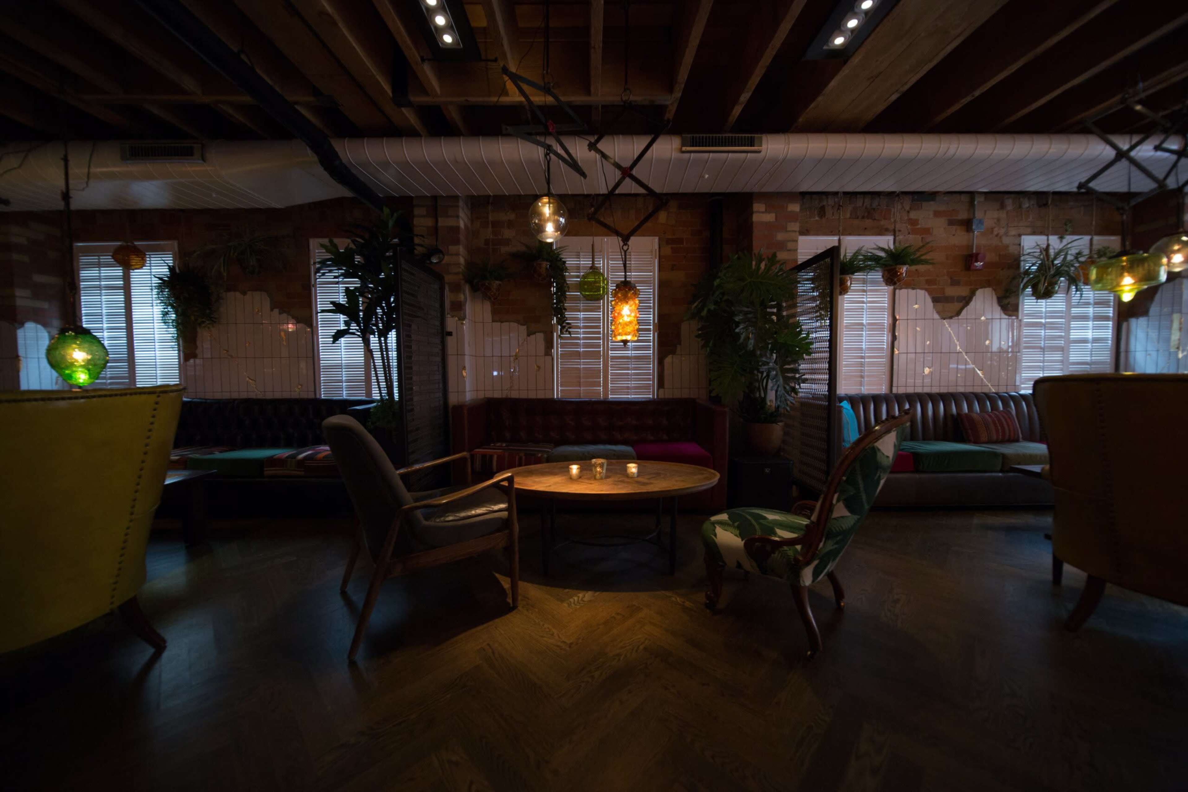 The image depicts a cozy lounge area with various seating options, soft lighting, and potted plants against a backdrop of brick walls and large windows.