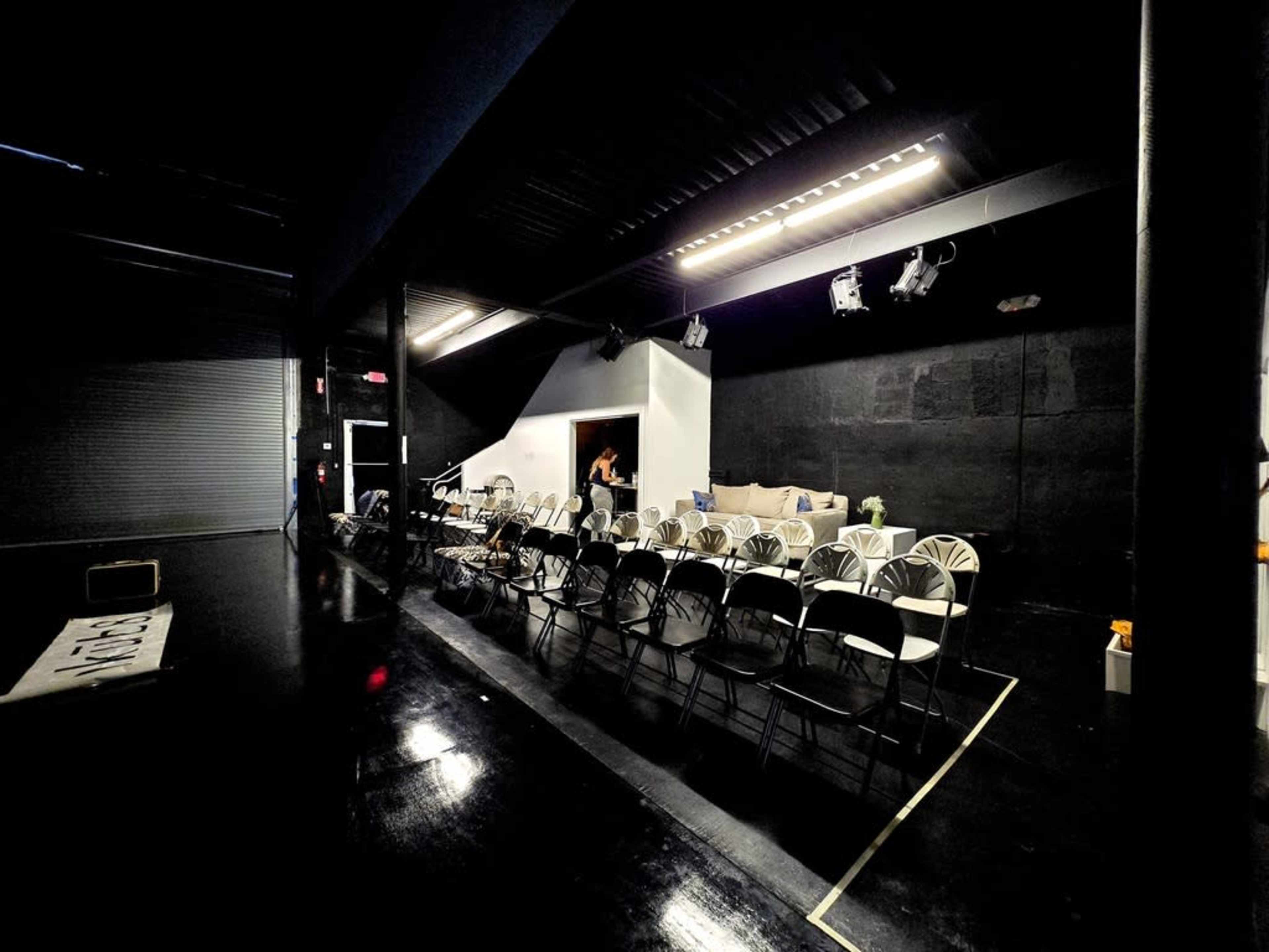 The image shows a dimly lit performance space with rows of folding chairs arranged in front of a white wall and a small stage area.