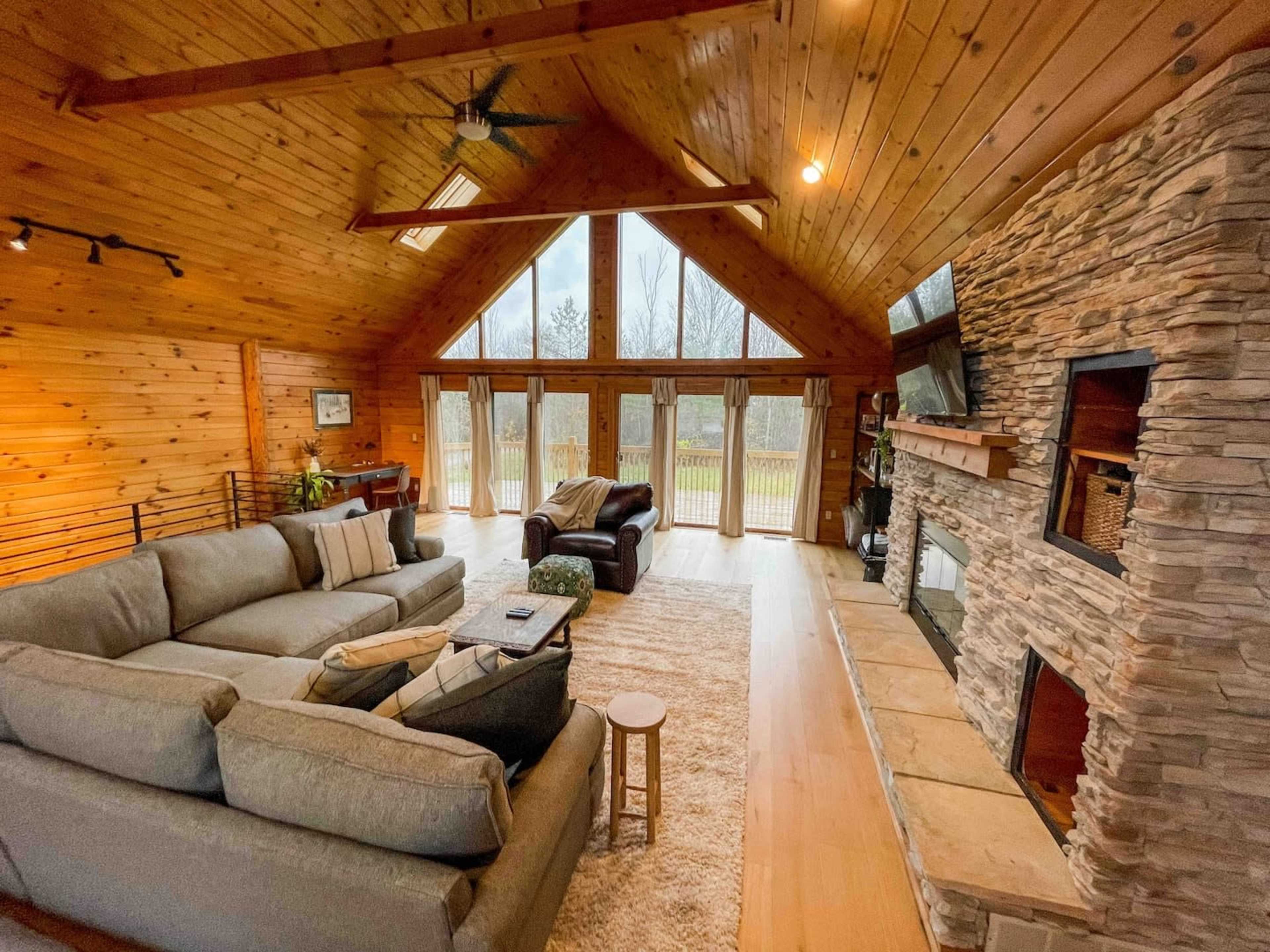 The living room features a large sectional sofa, a stone fireplace, and large windows framed by wooden walls, allowing natural light to illuminate the space.