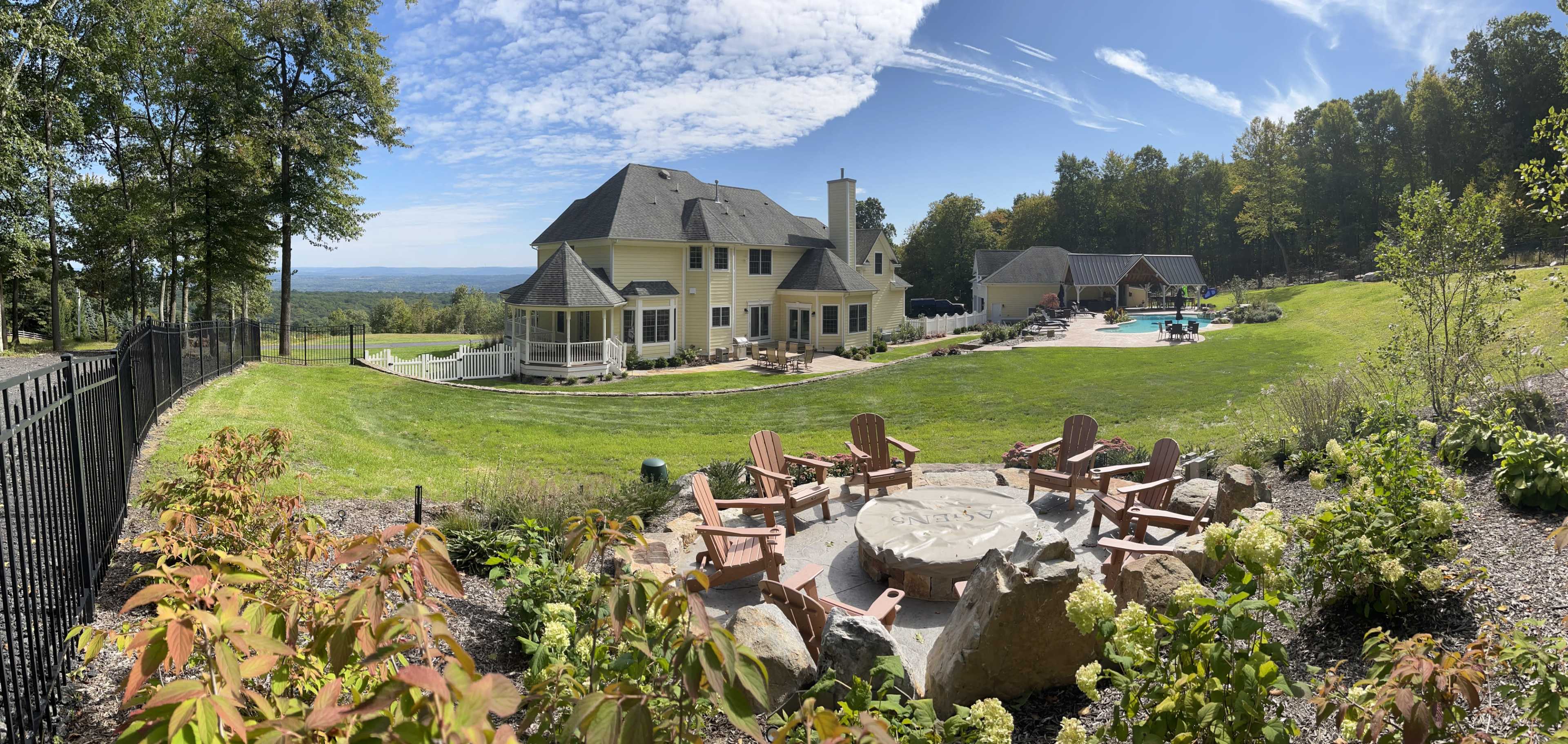A large yellow house with multiple sections overlooks a spacious green lawn, a stone fire pit surrounded by chairs, and a pool area, all set against a backdrop of trees and blue sky.