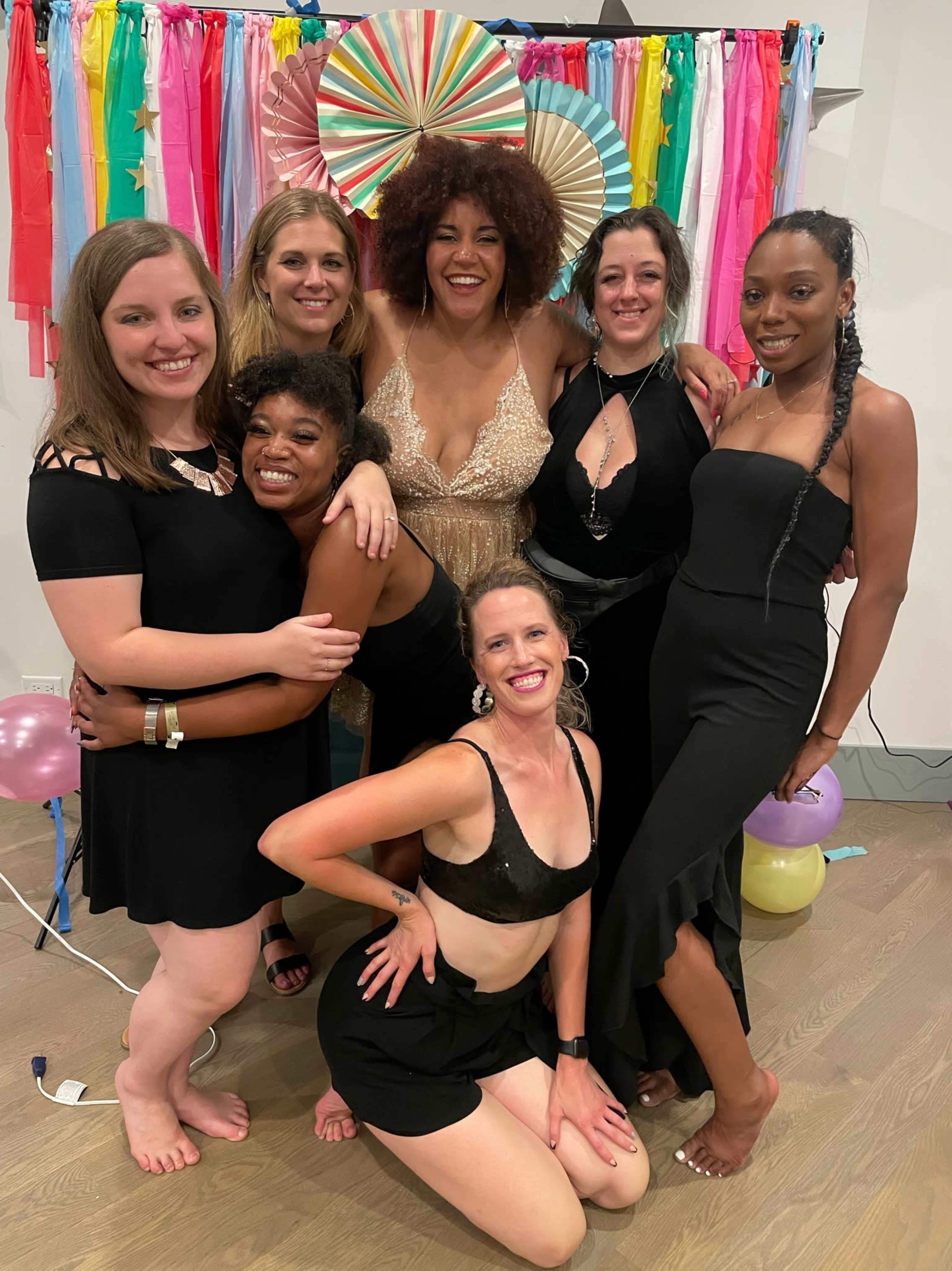 A group of six women poses together, wearing black dresses and celebrating in front of a colorful backdrop.
