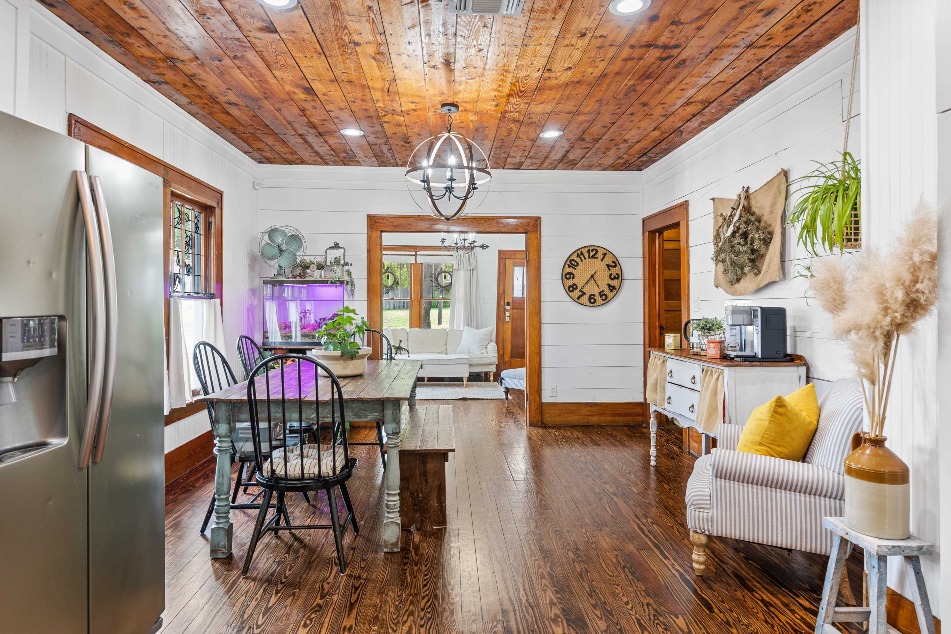 The image shows a bright, rustic kitchen and dining area featuring a wooden ceiling, a large dining table, and a mix of modern and vintage decor.