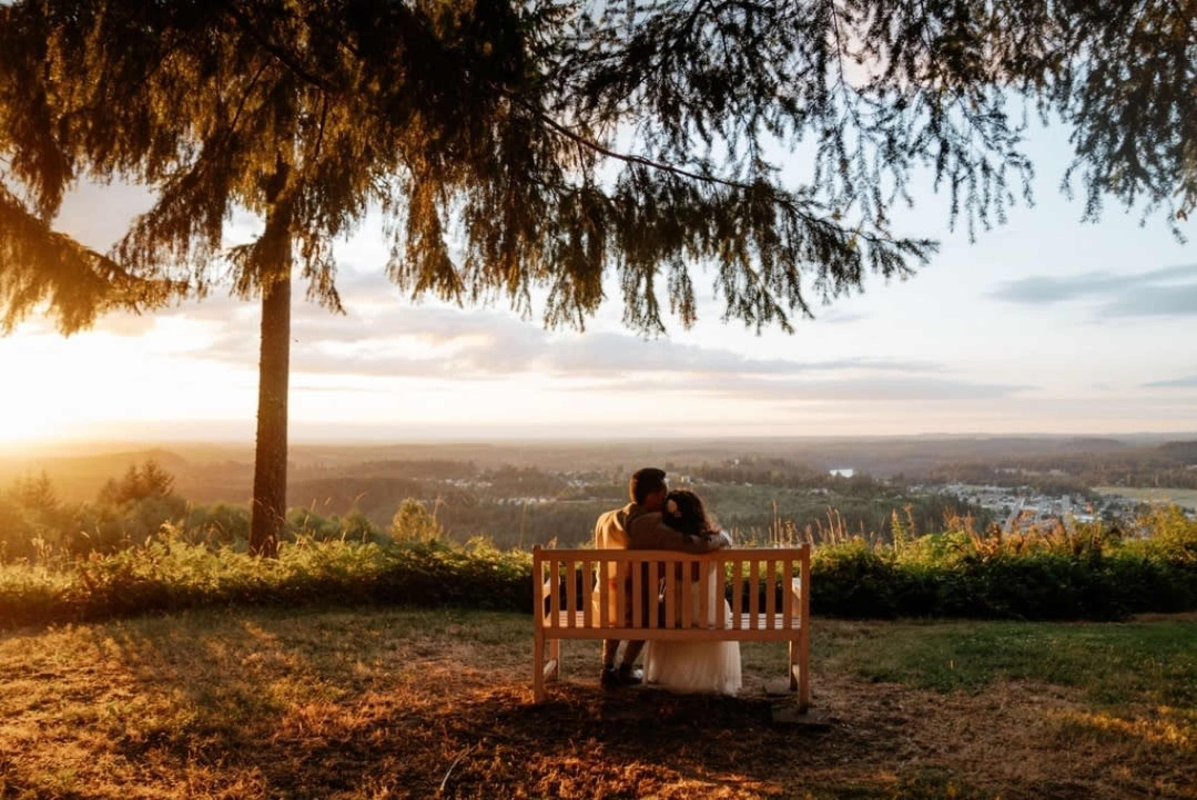 A couple sits closely on a wooden bench overlooking a valley at sunset, framed by tall trees.