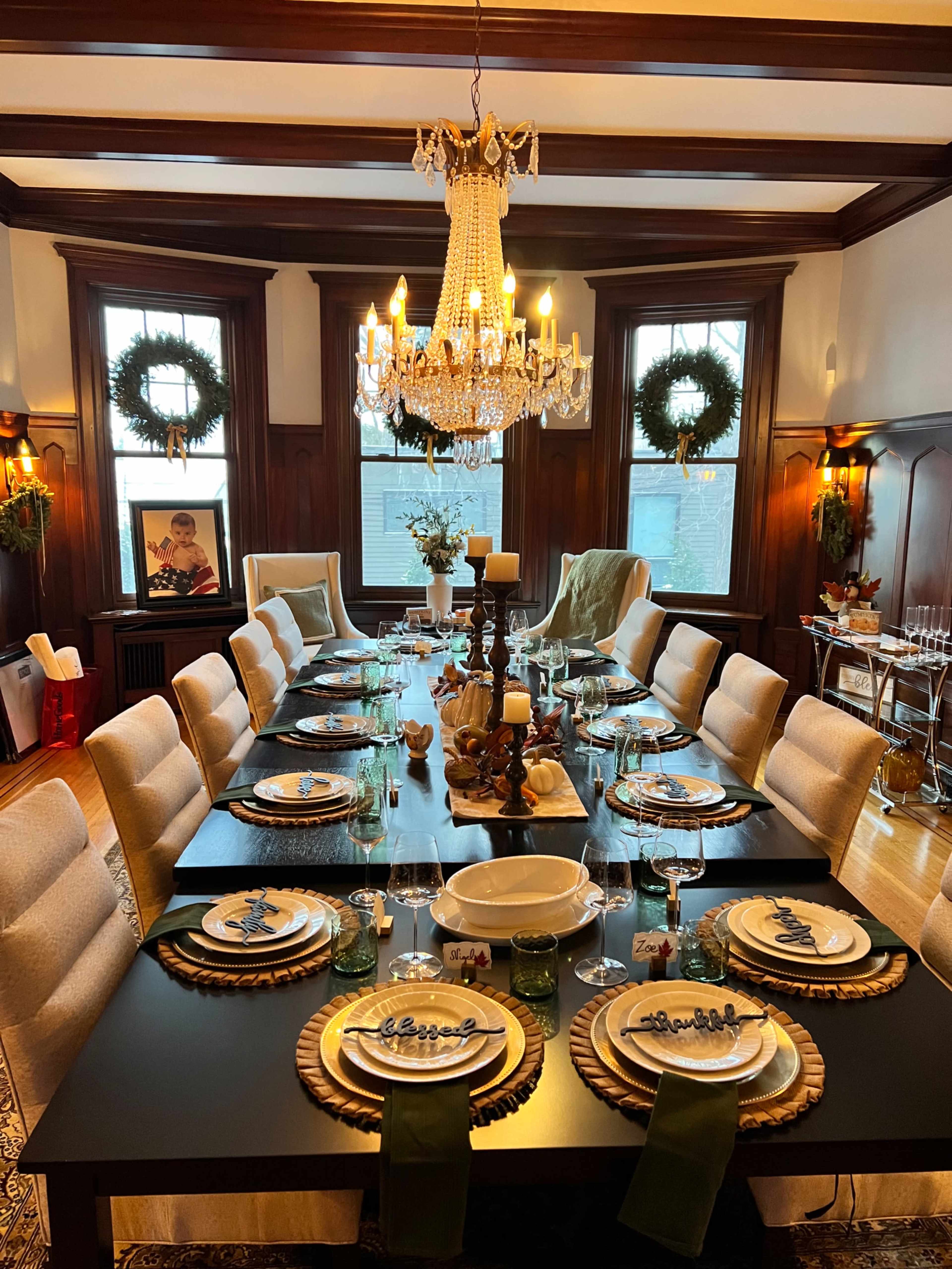 A formal dining room is set with an elaborate table arrangement featuring elegant place settings, a central candle display, and festive wreaths on the windows.