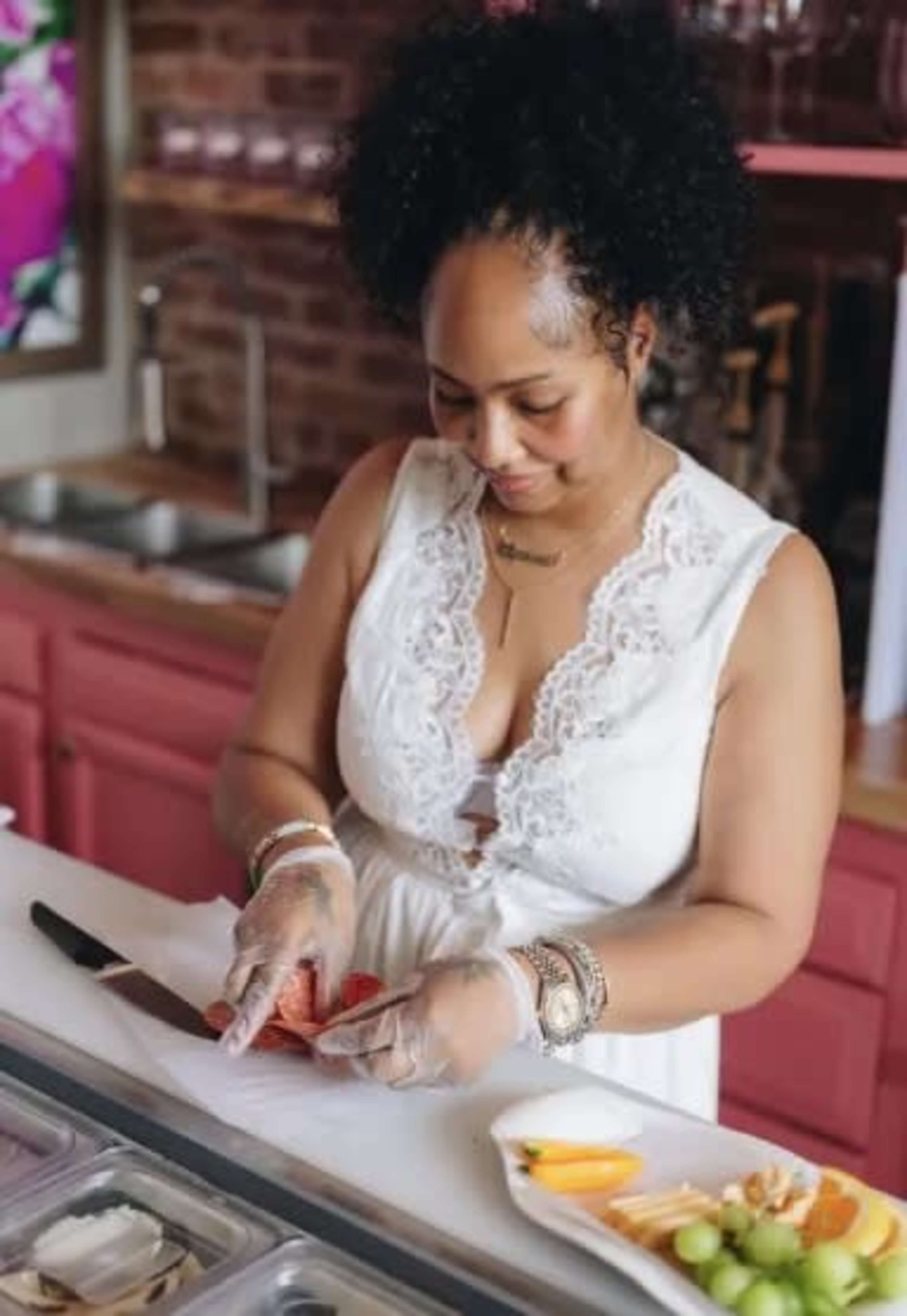A woman with curly hair is cutting fruit at a counter while wearing gloves and a white dress.