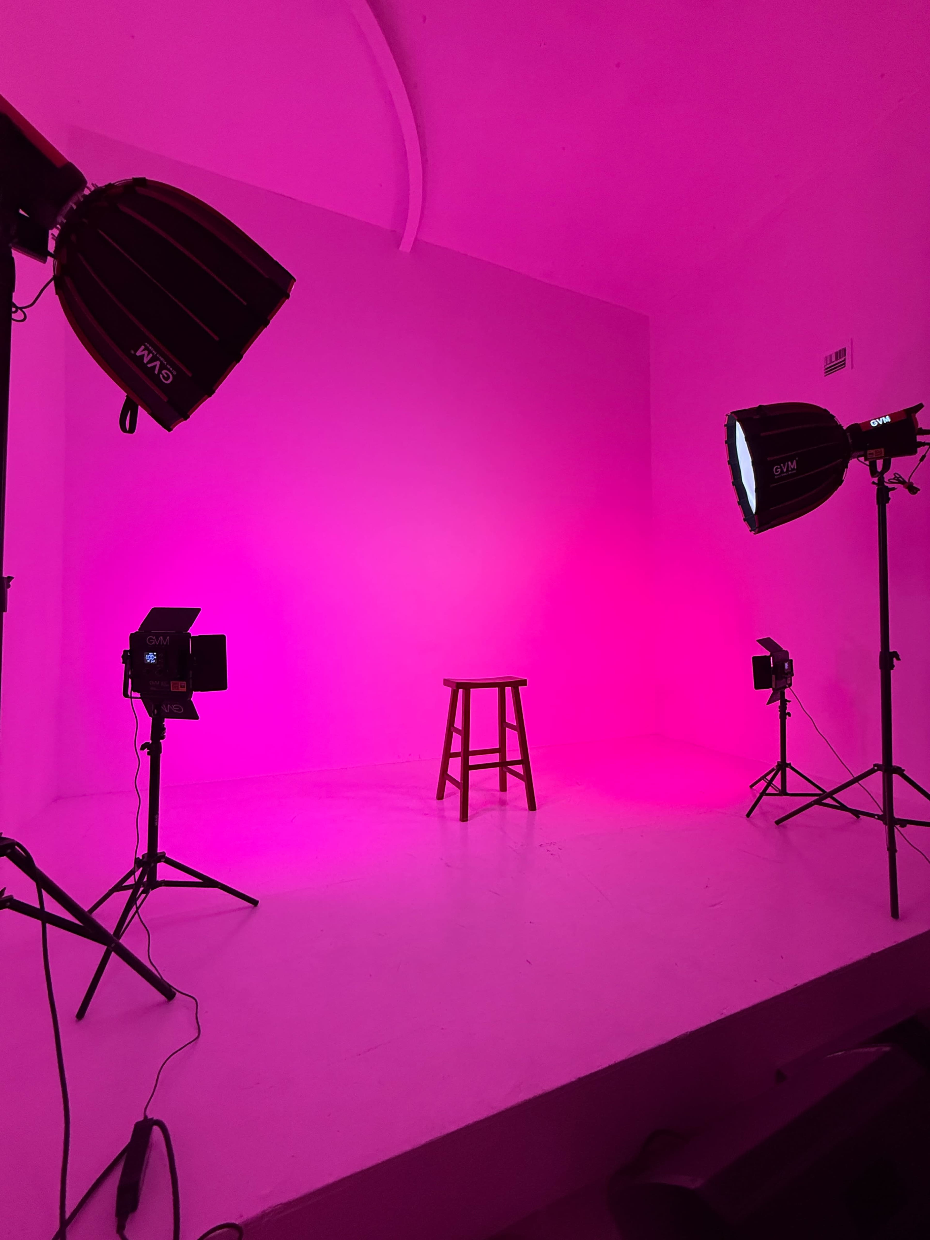 A wooden stool is centered in an empty studio illuminated by pink lighting from various studio lights.