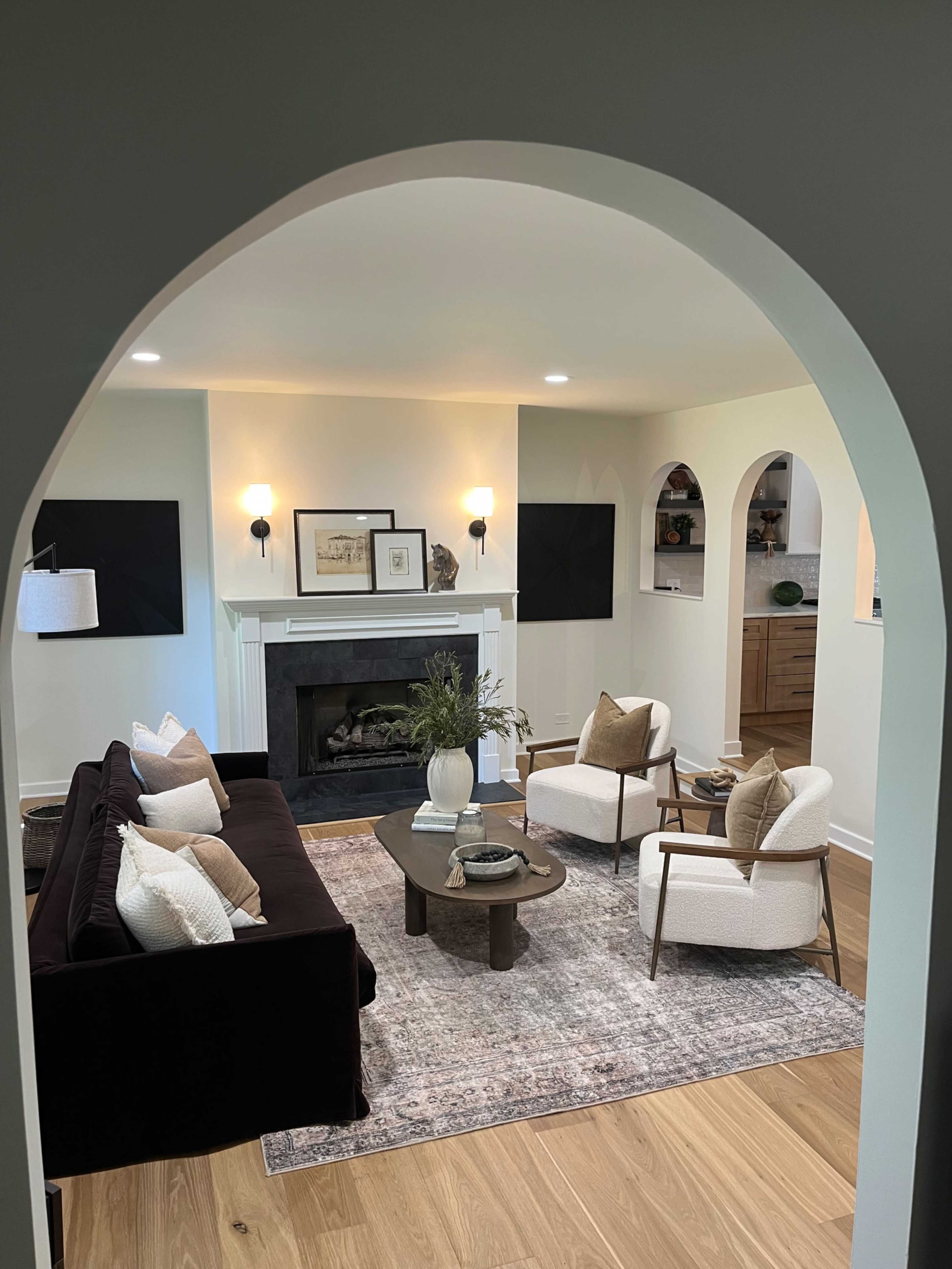 The image shows a modern living room featuring a dark sofa, two light-colored armchairs, a coffee table, and a fireplace, framed by an arched entryway.