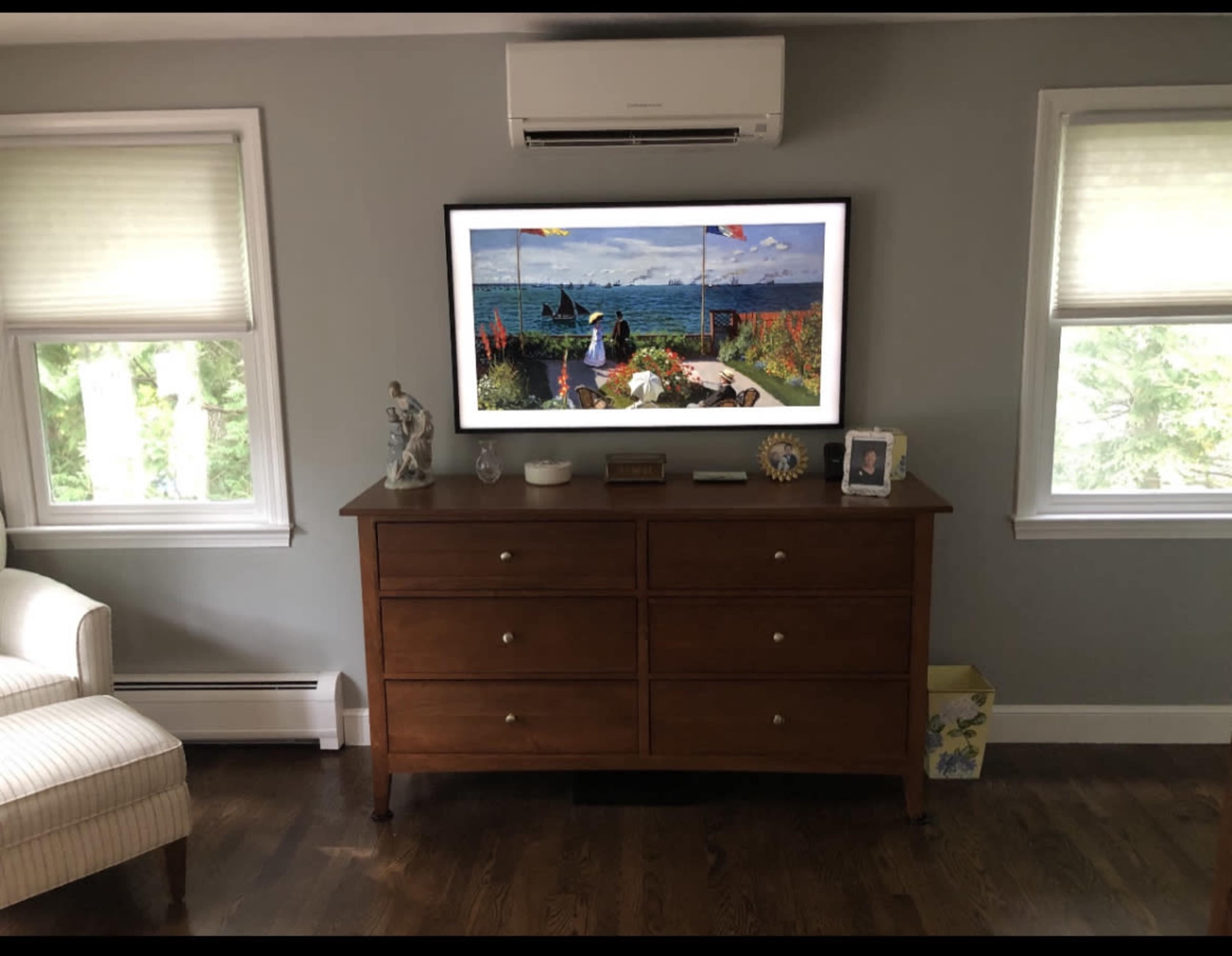 A living room with a wooden dresser under a wall-mounted TV displaying a scenic landscape, accompanied by a small decorative statue and a framed photo on the dresser.