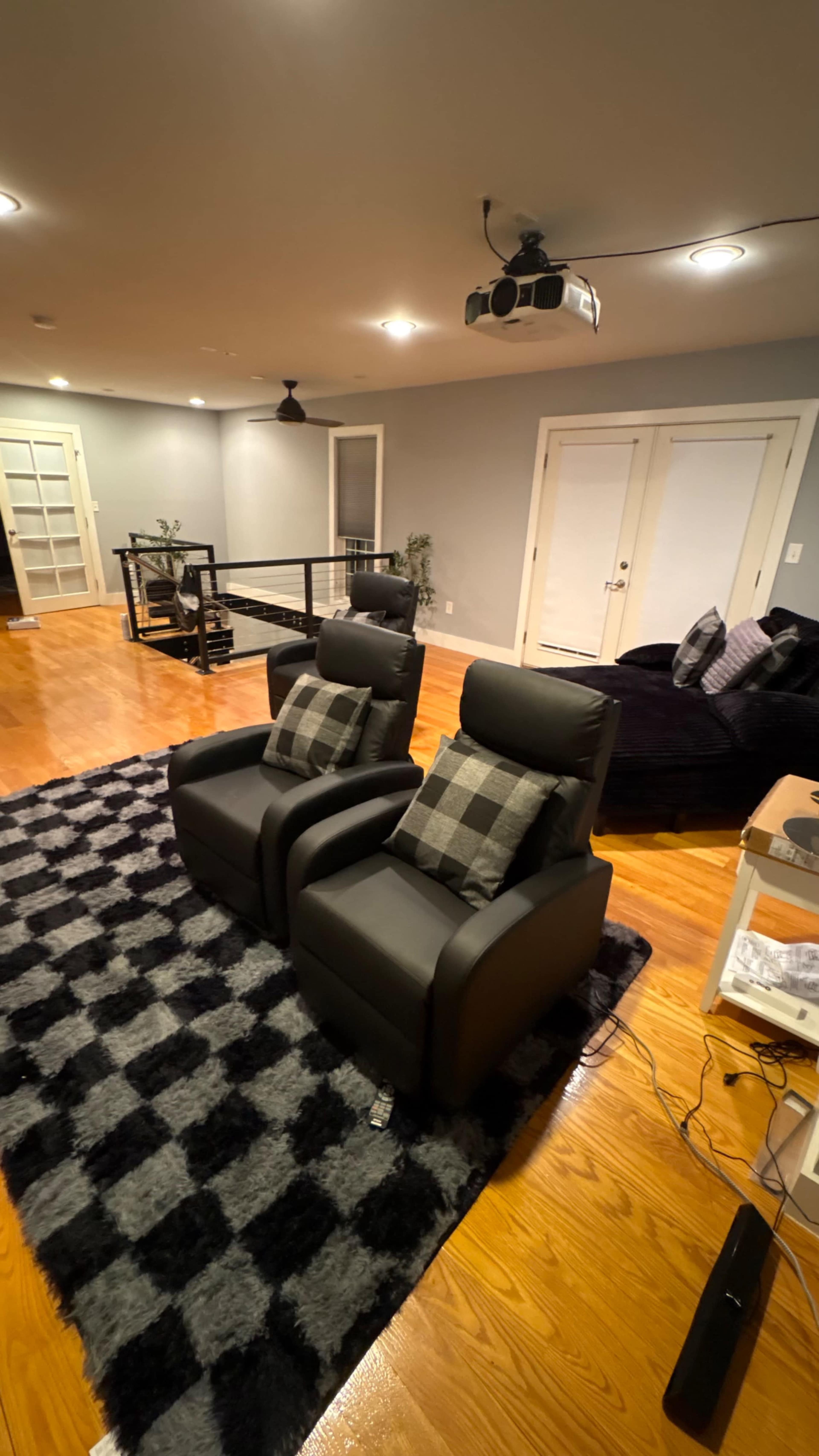 The image shows a spacious living area with two black reclining chairs, a checkered rug, hardwood flooring, and a projector mounted on the ceiling.