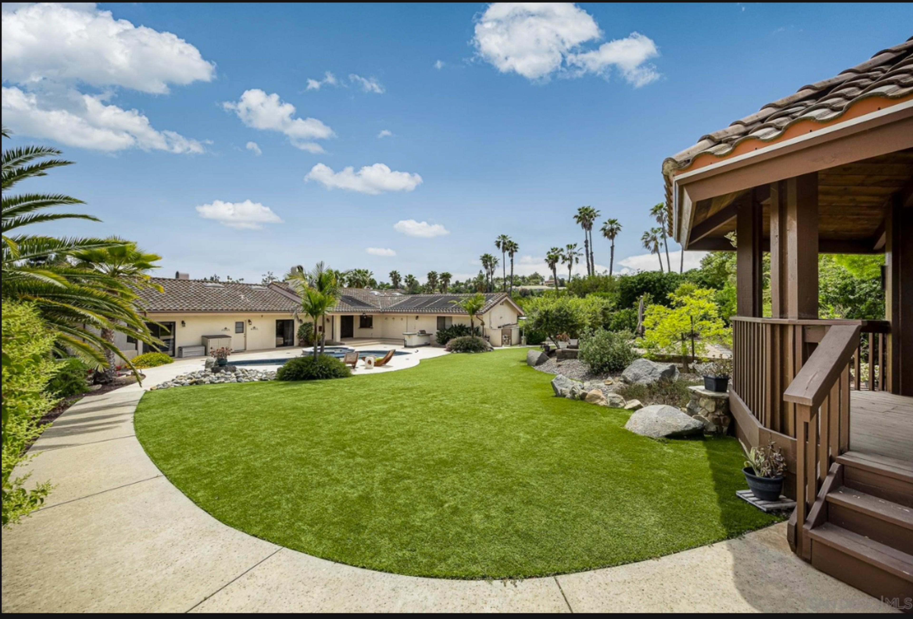 The image shows a spacious backyard featuring a pool, landscaped greenery, and a patio area surrounded by palm trees and boulders.