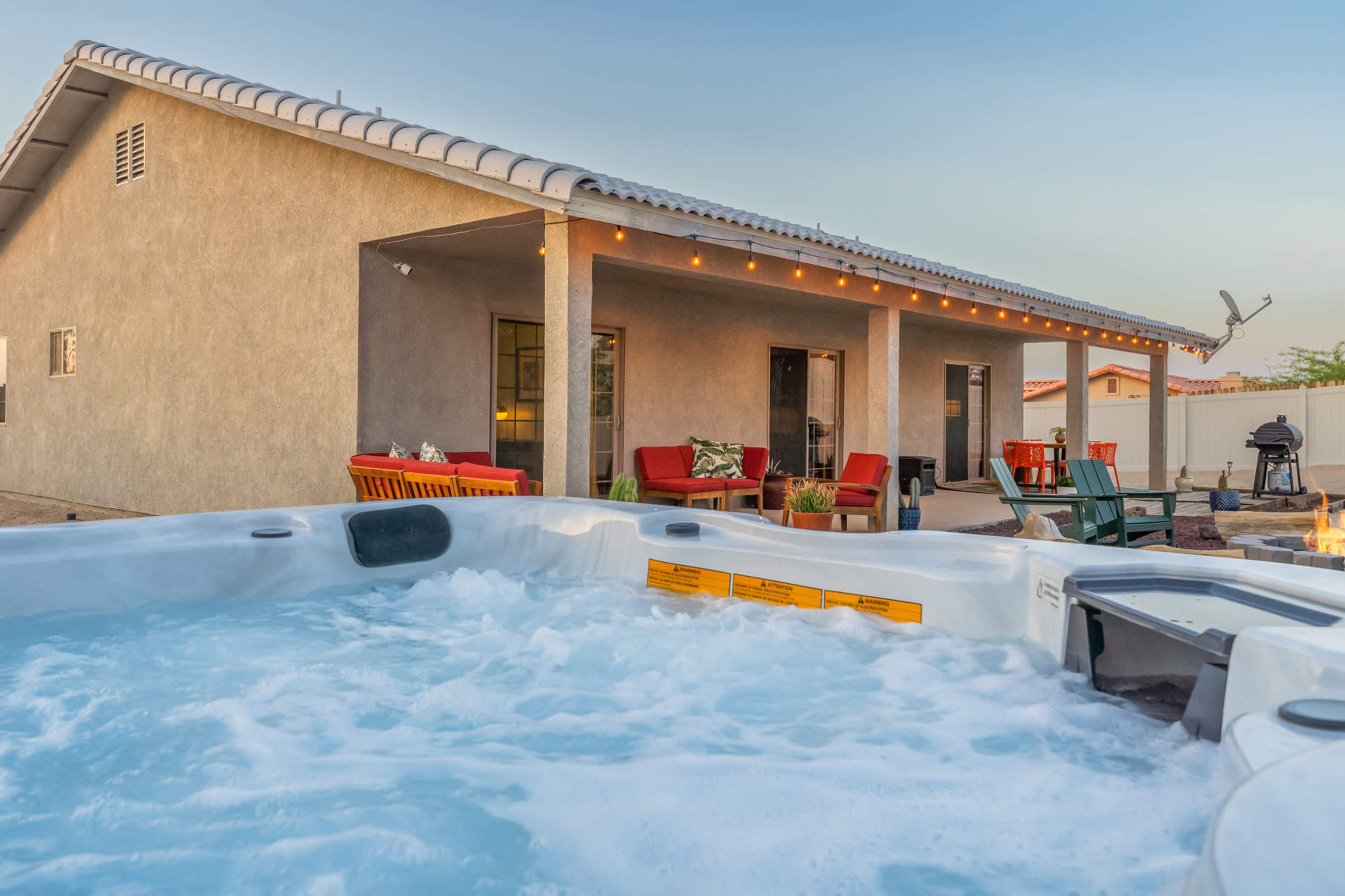 A hot tub in the foreground with a backyard patio area featuring seating and string lights attached to a nearby house.