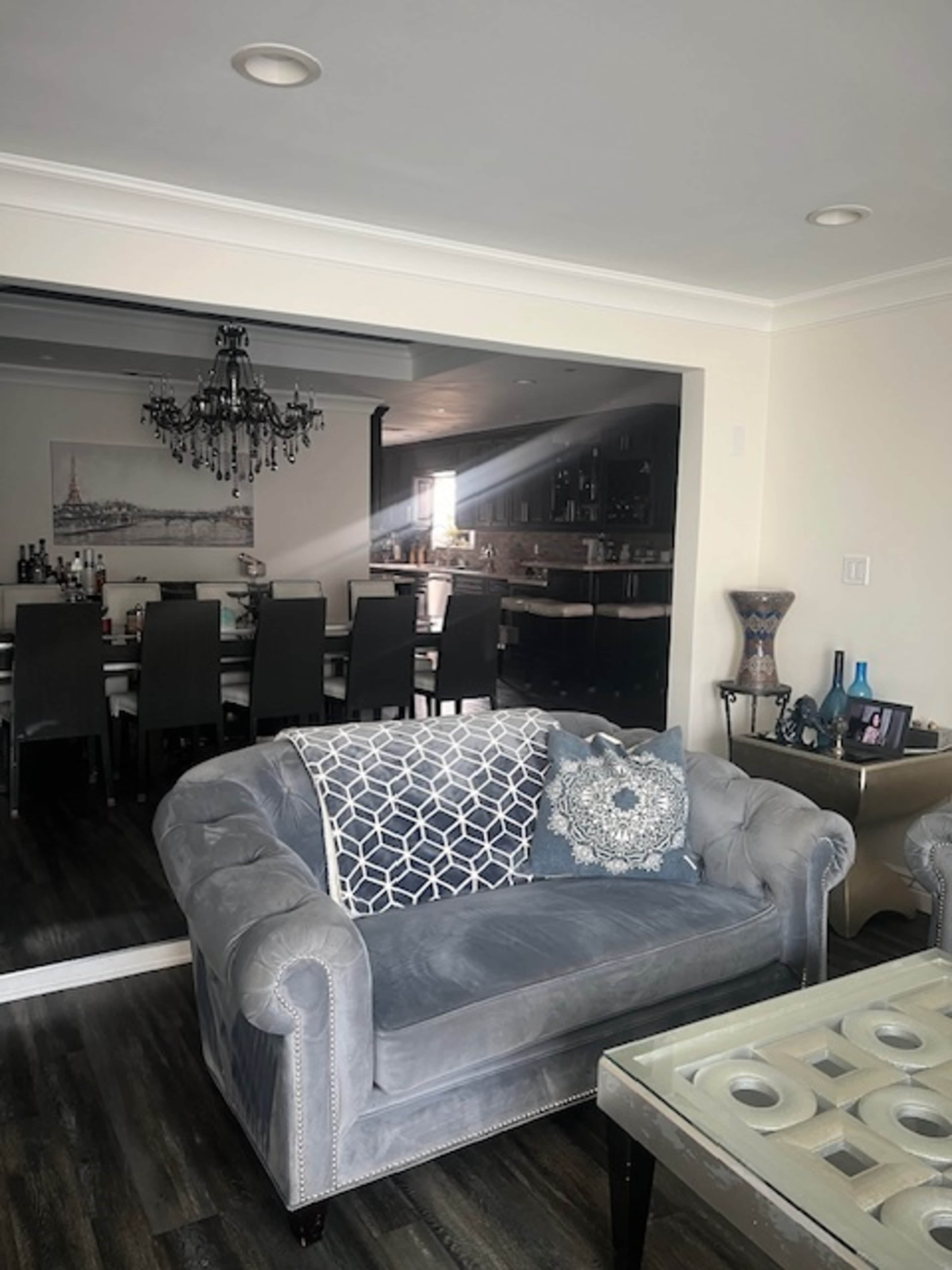 A gray velvet couch with decorative pillows is positioned in a well-lit living room that opens to a dining area with a large table and dark cabinetry.